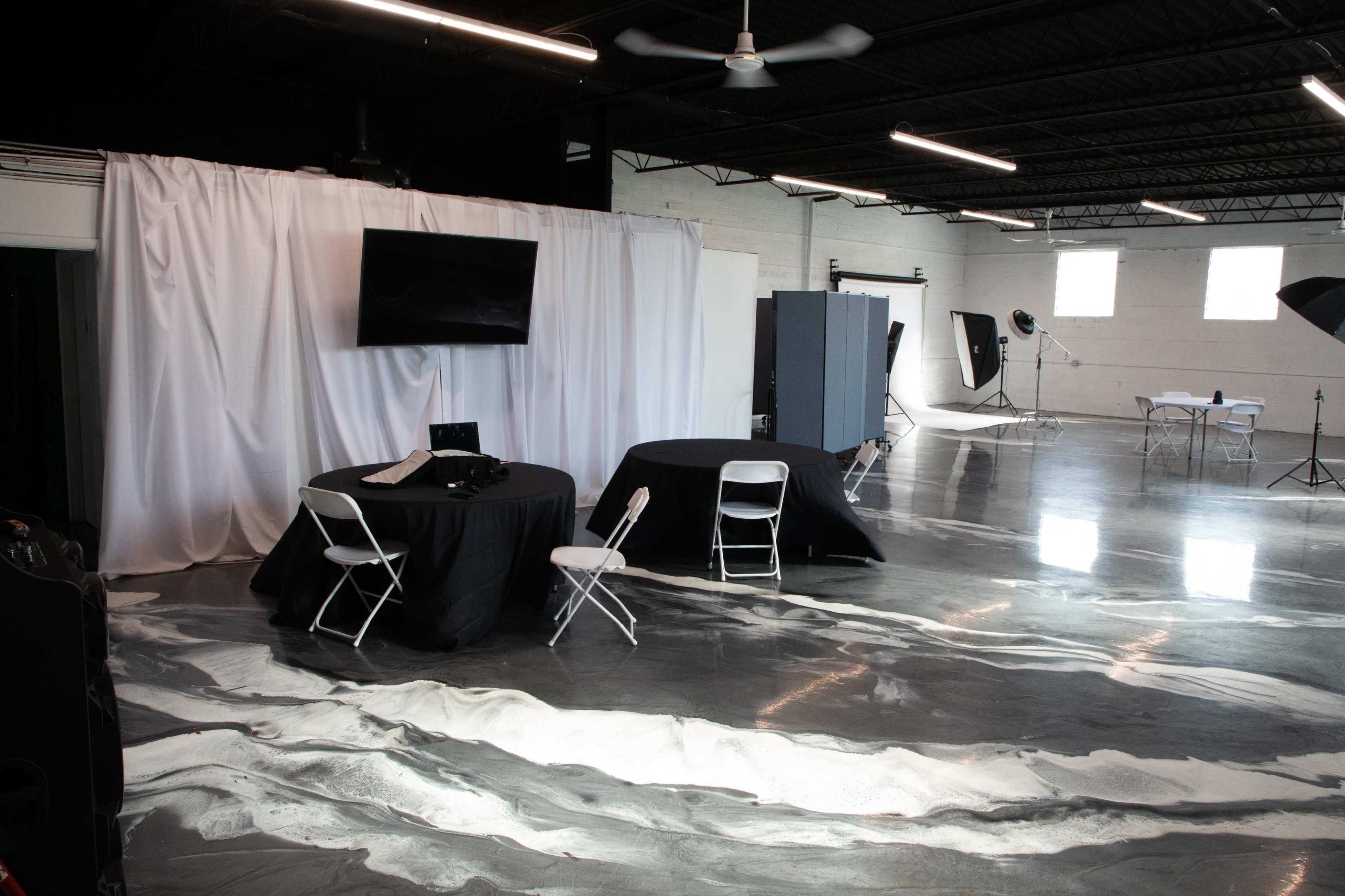 A photography studio features a polished concrete floor with a marble-like design, two tables dressed in black cloth, and various lighting equipment set up around the space.