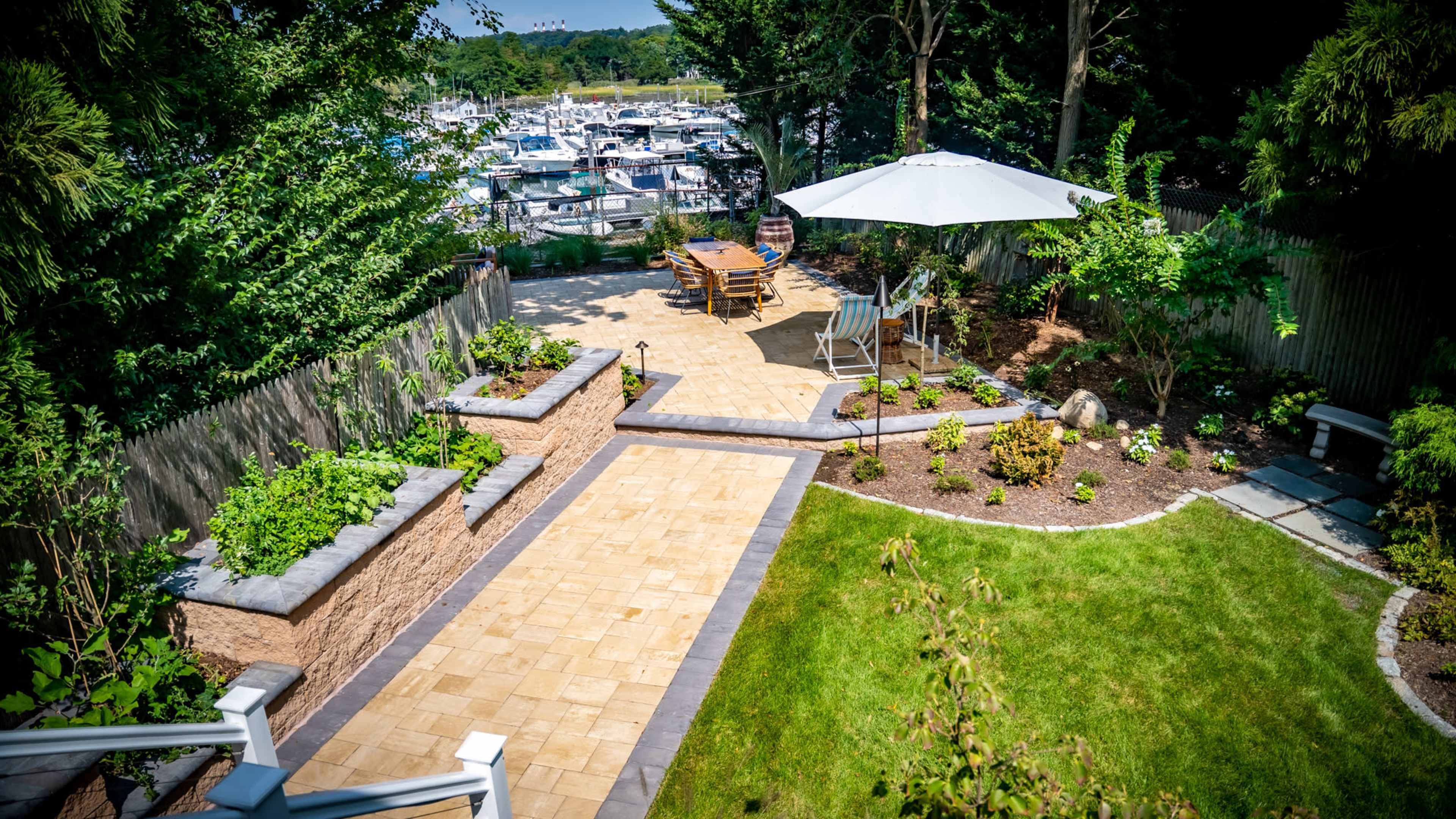 The image shows a landscaped outdoor patio with seating, surrounded by plants and leading to a marina in the background.