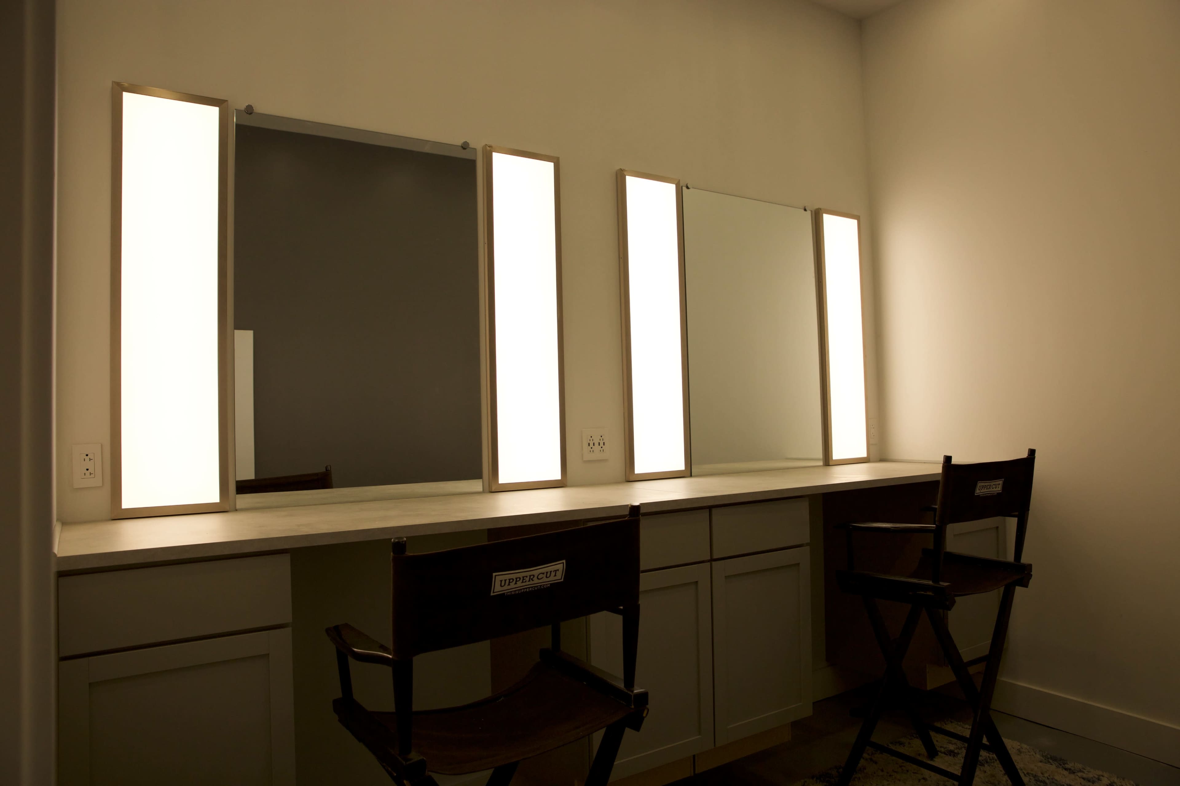 A well-lit vanity area with two mirrors and two director's chairs against a plain wall.
