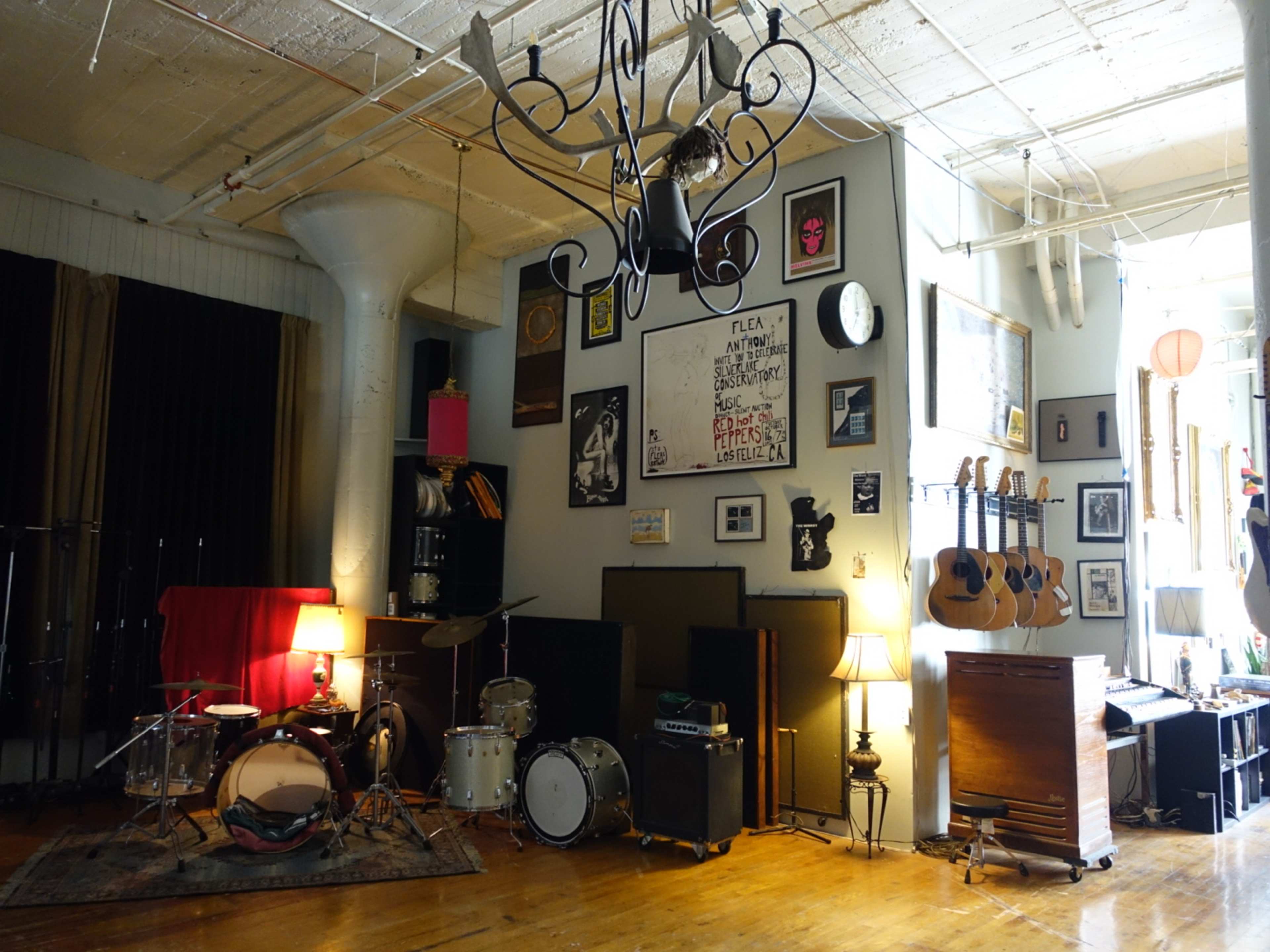 The image shows a cozy music studio with a drum set, various guitars hanging on the wall, and framed artwork alongside a vintage piano in a well-lit space.