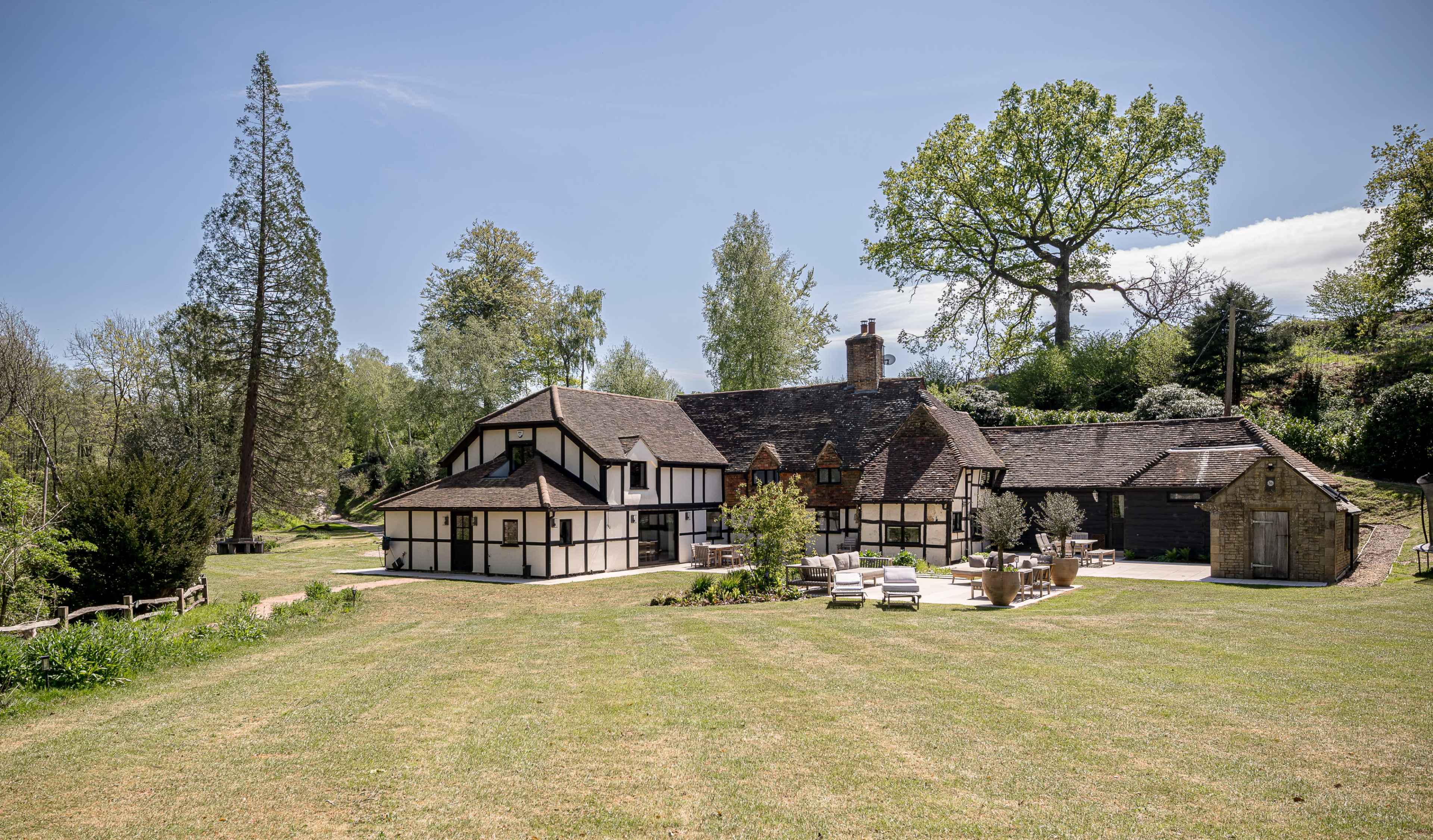 A large, traditional-style house with a mix of timber framing and stone accents is surrounded by a well-maintained lawn and trees.