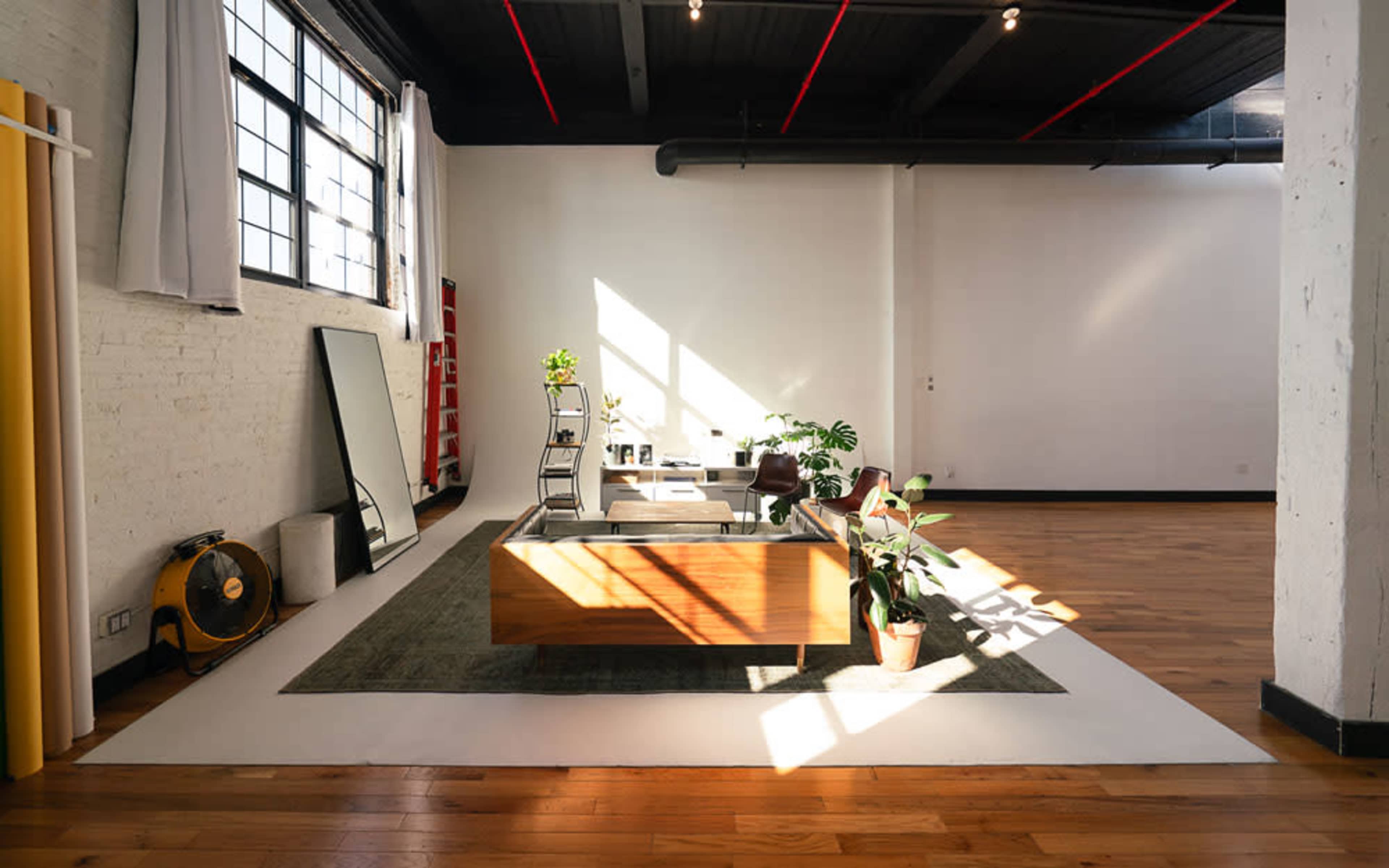The image shows a spacious, modern office interior with a wooden coffee table surrounded by plants and a large window allowing natural light to illuminate the room.