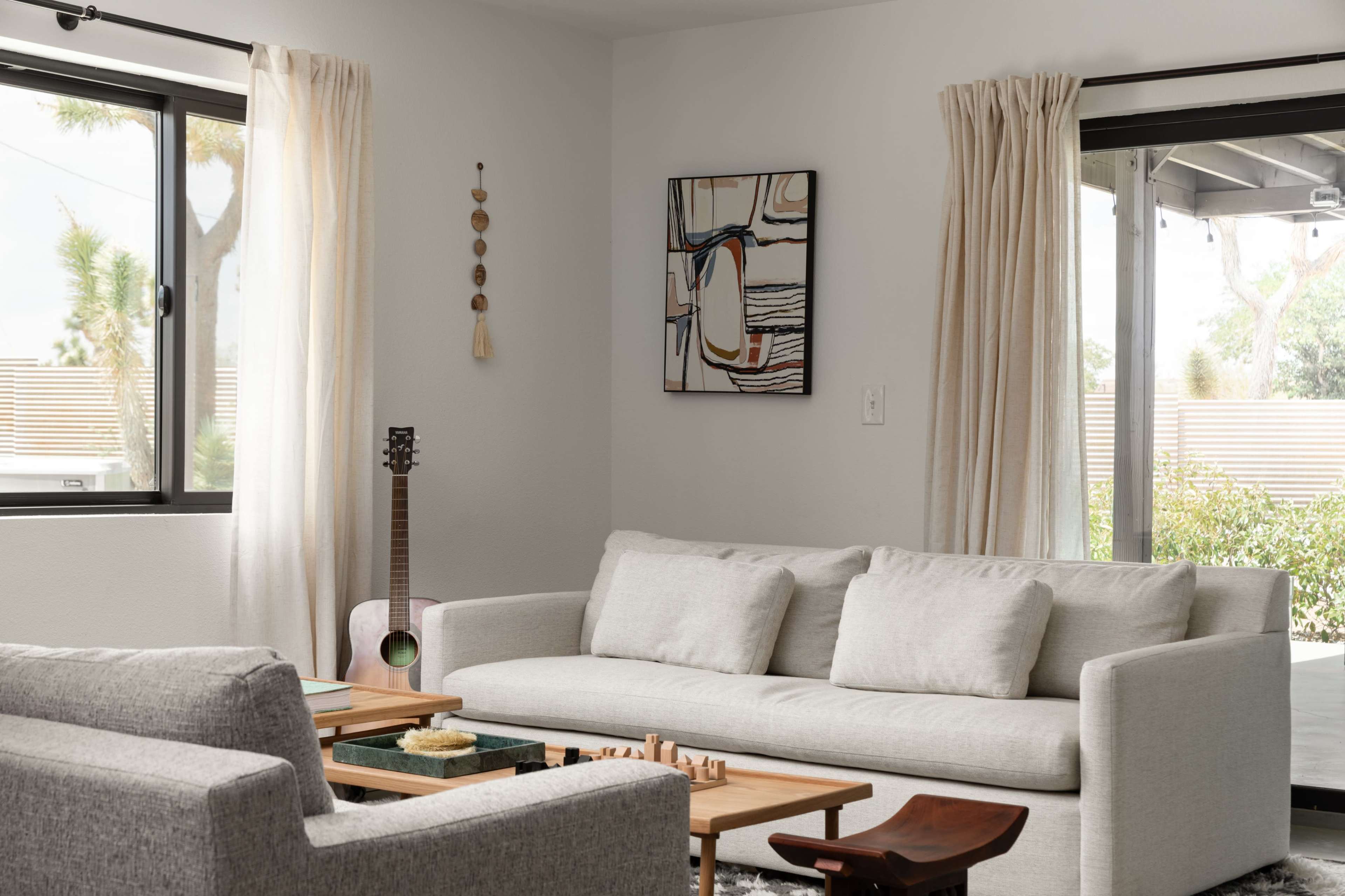 The image shows a modern living room with two light-colored sofas, a wooden coffee table with a chess set, and large windows allowing natural light to enter.