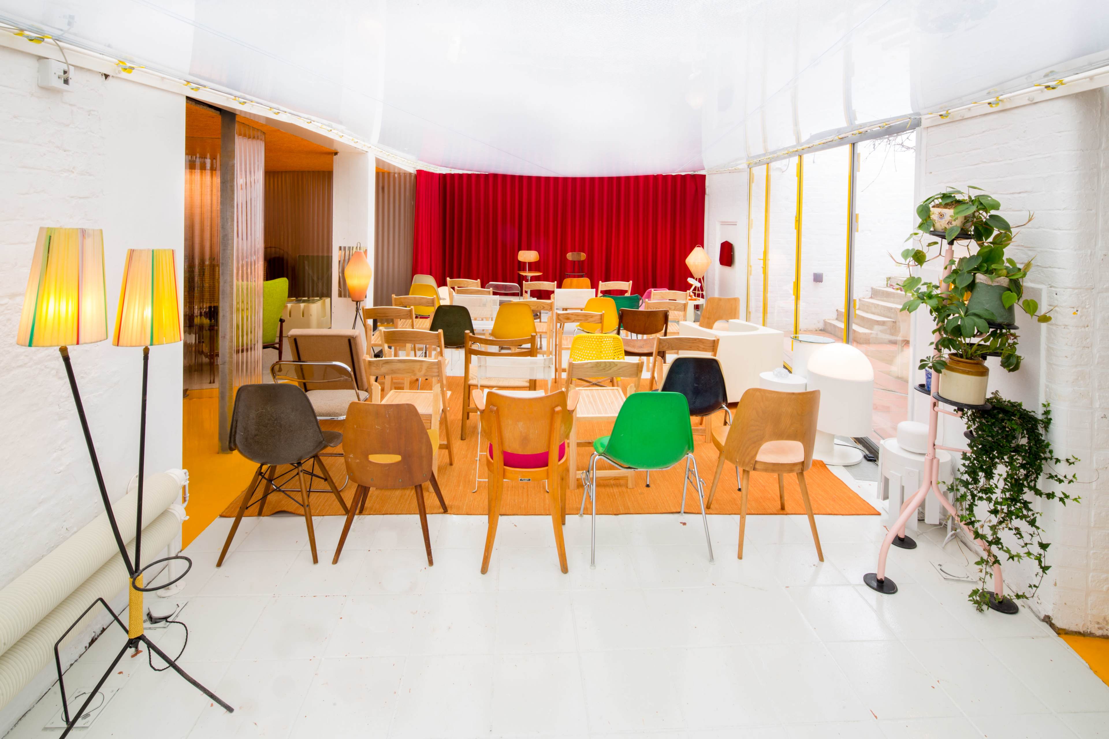 The image shows a brightly lit room with various chairs arranged in rows facing a red curtain, with decorative lamps and plants present in the space.
