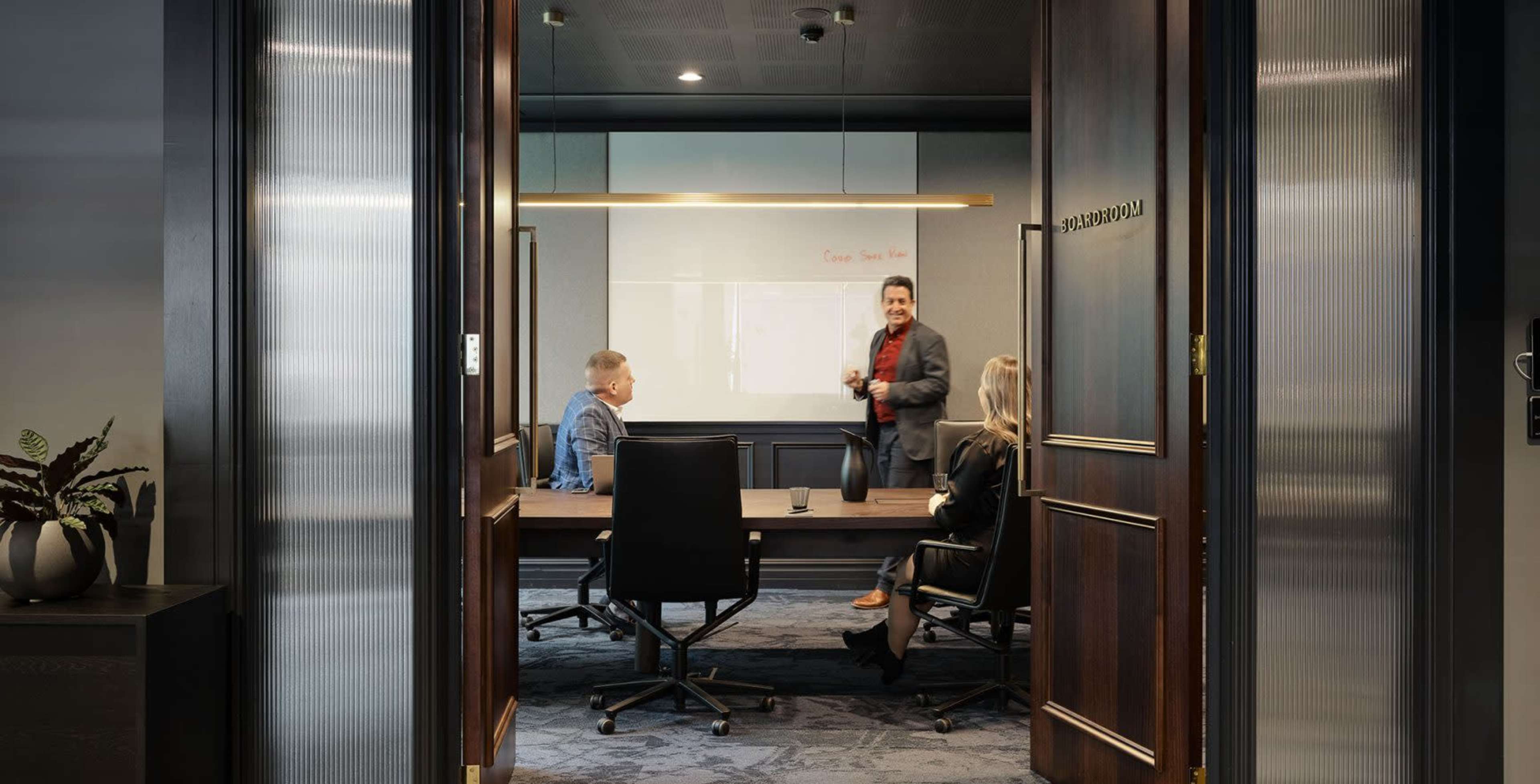 A man presents to two colleagues in a conference room with a large screen displaying information.