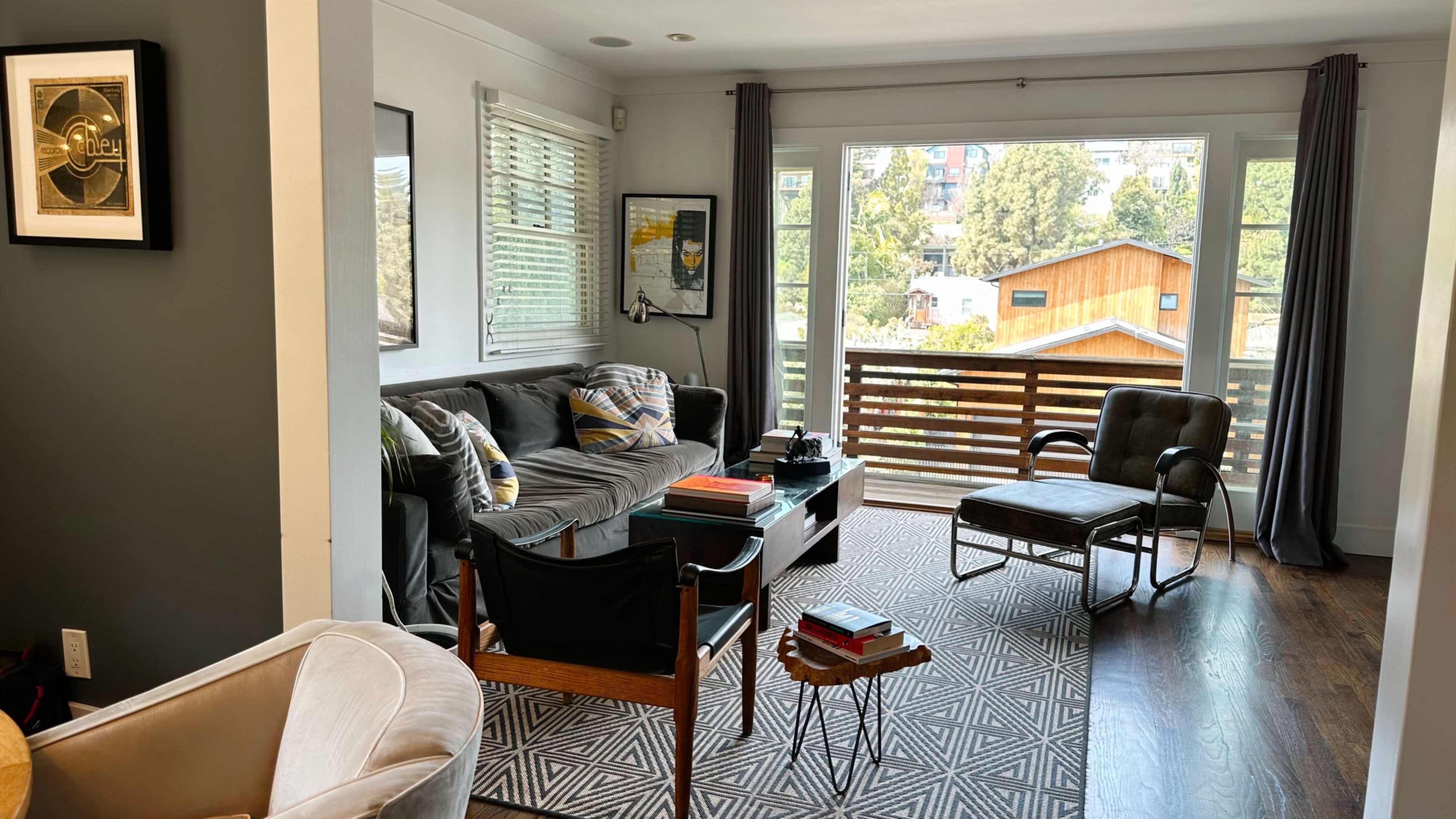 The image shows a living room with a gray sofa, two armchairs, a patterned rug, and large windows overlooking a view of greenery and houses.
