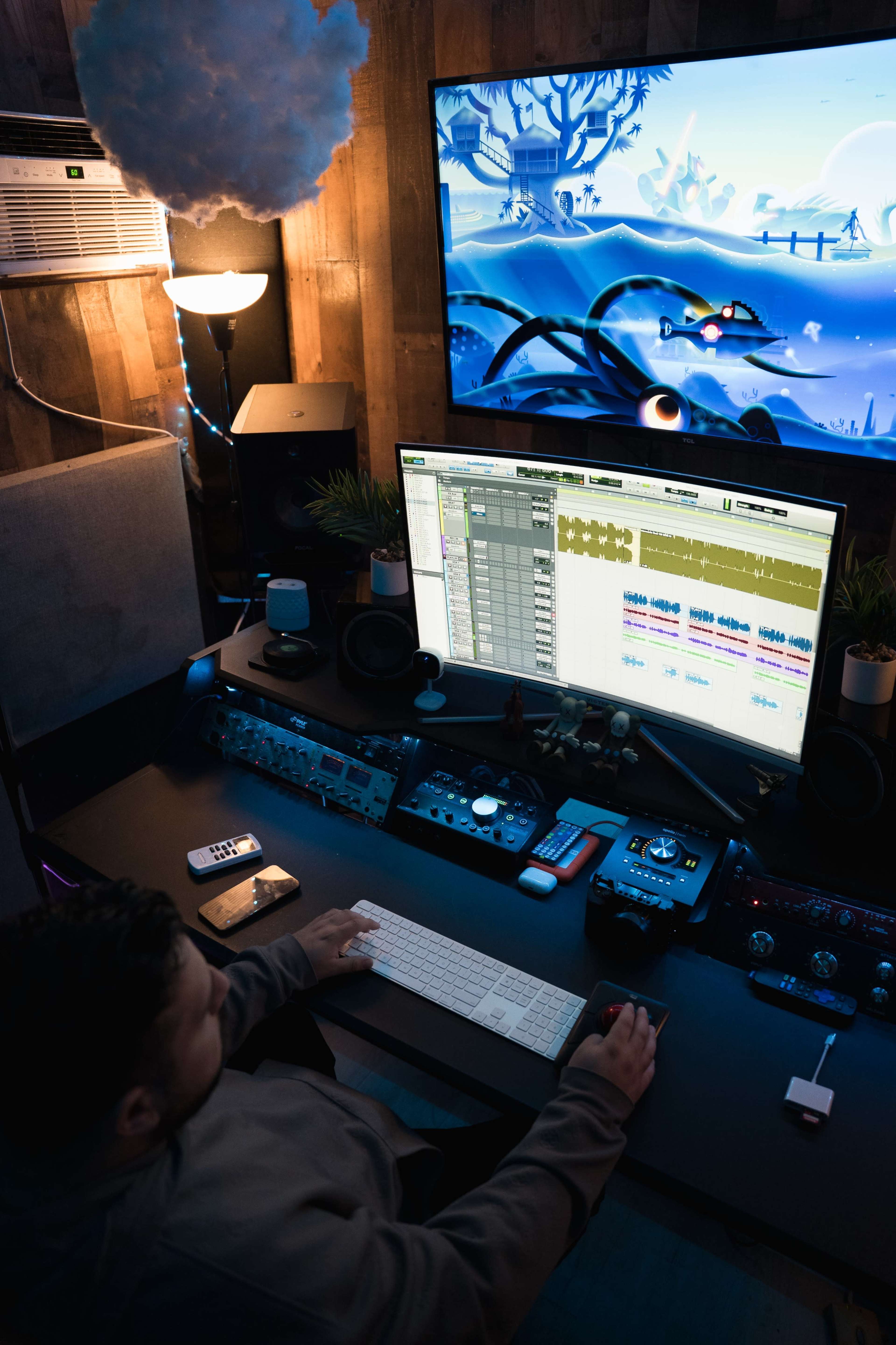 A person is sitting at a desk with a large computer monitor displaying animated artwork, surrounded by audio equipment and a cozy room setup.