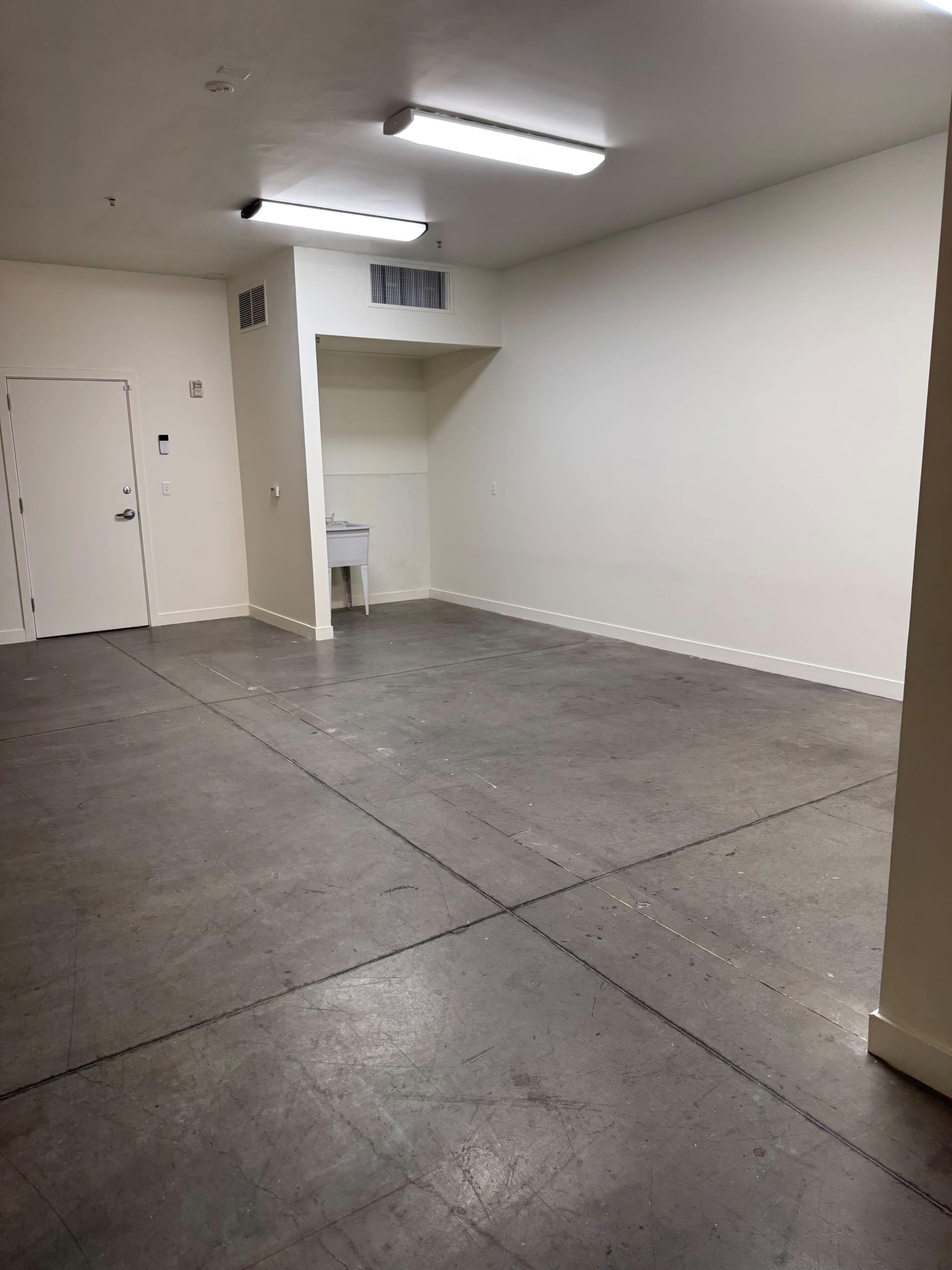 The image shows a spacious, vacant room with concrete flooring, white walls, and a small utility area in the corner with a sink.