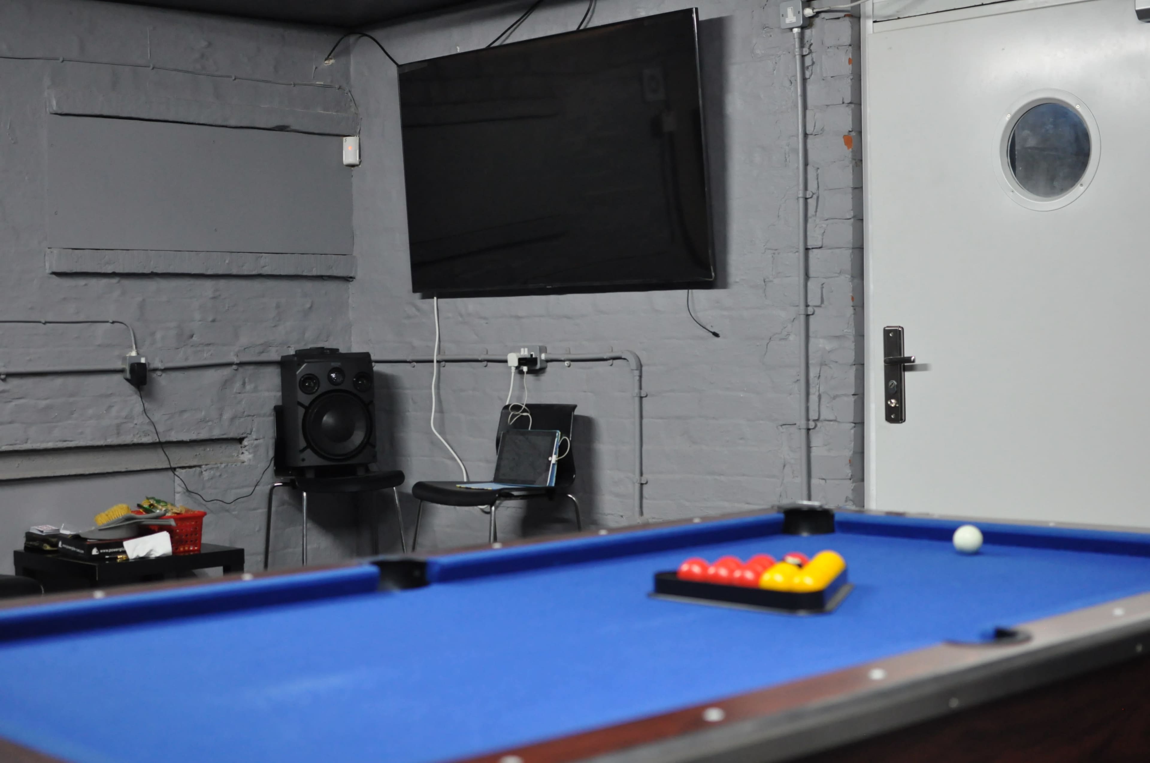 A blue pool table is set up with a rack of colorful balls in a dimly lit room featuring a flat-screen TV and a doorway.