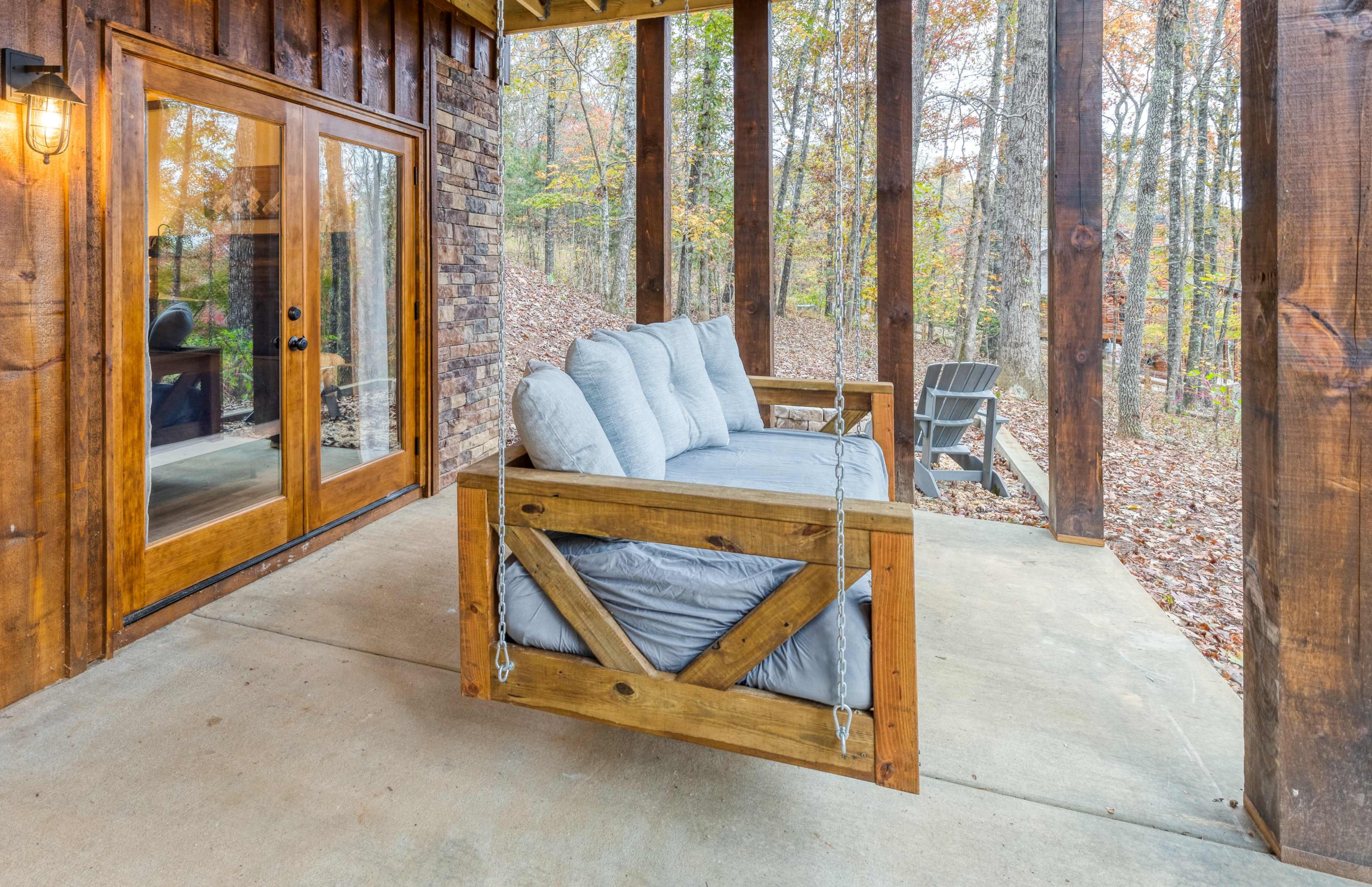 A wooden porch features a suspended bench and large windows overlooking a forested area.