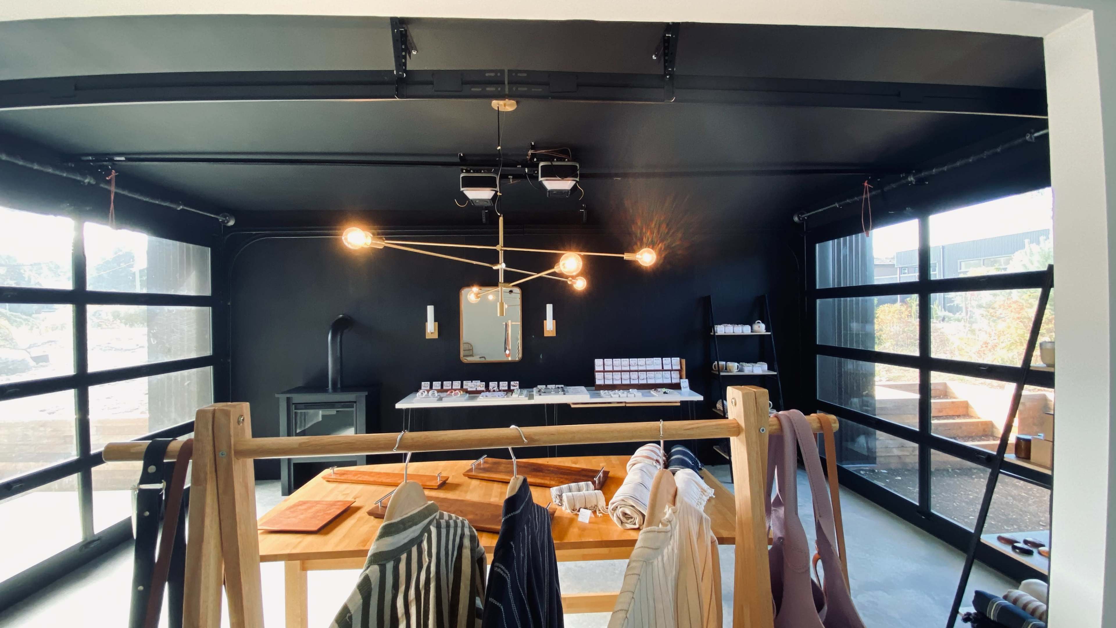 The image shows a modern, minimalist interior with a dark-colored wall, an industrial-style light fixture, and clothing hanging on a wooden rack in front of large glass doors.