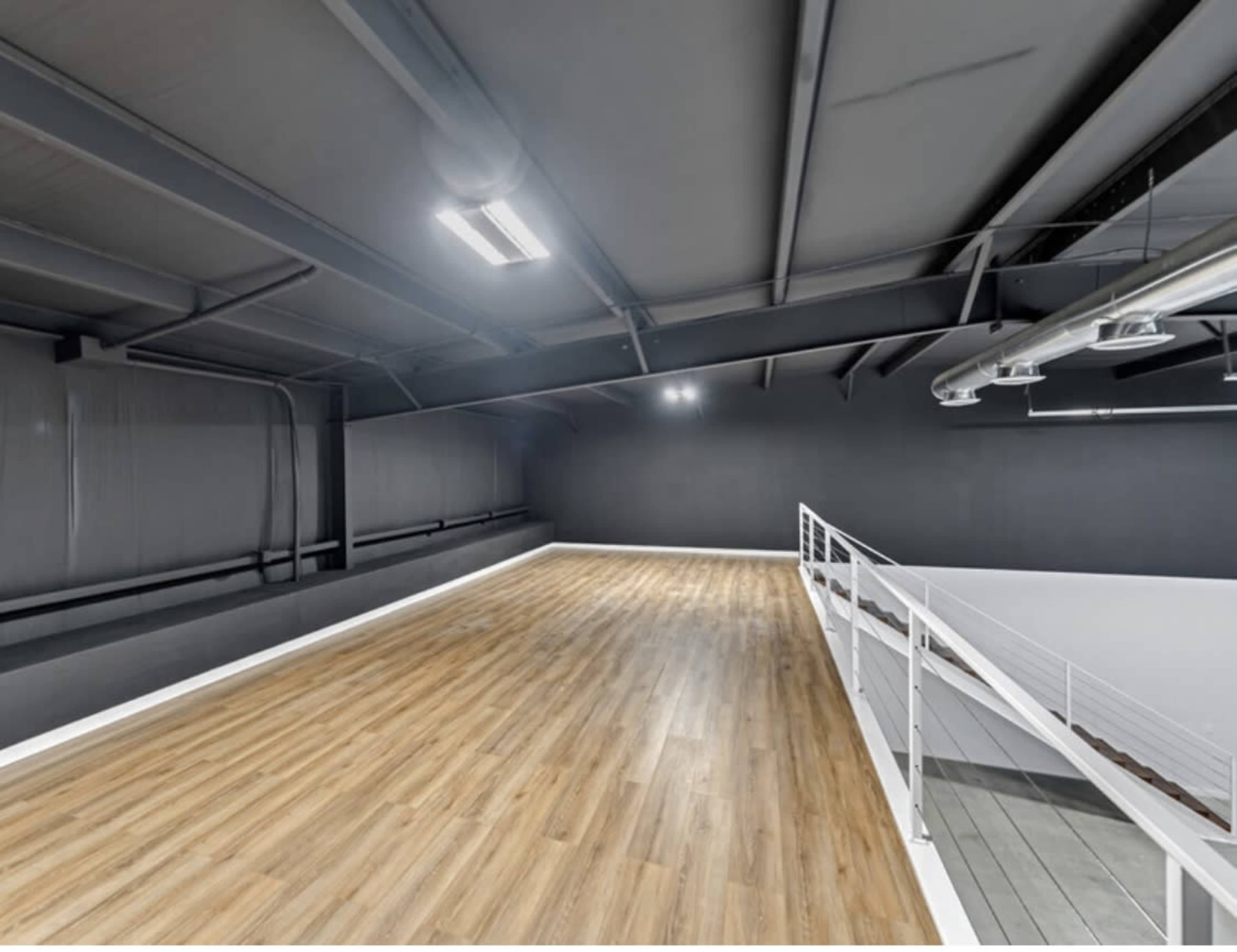 The image shows an empty, well-lit interior space with wooden flooring and a high ceiling supported by metal beams.