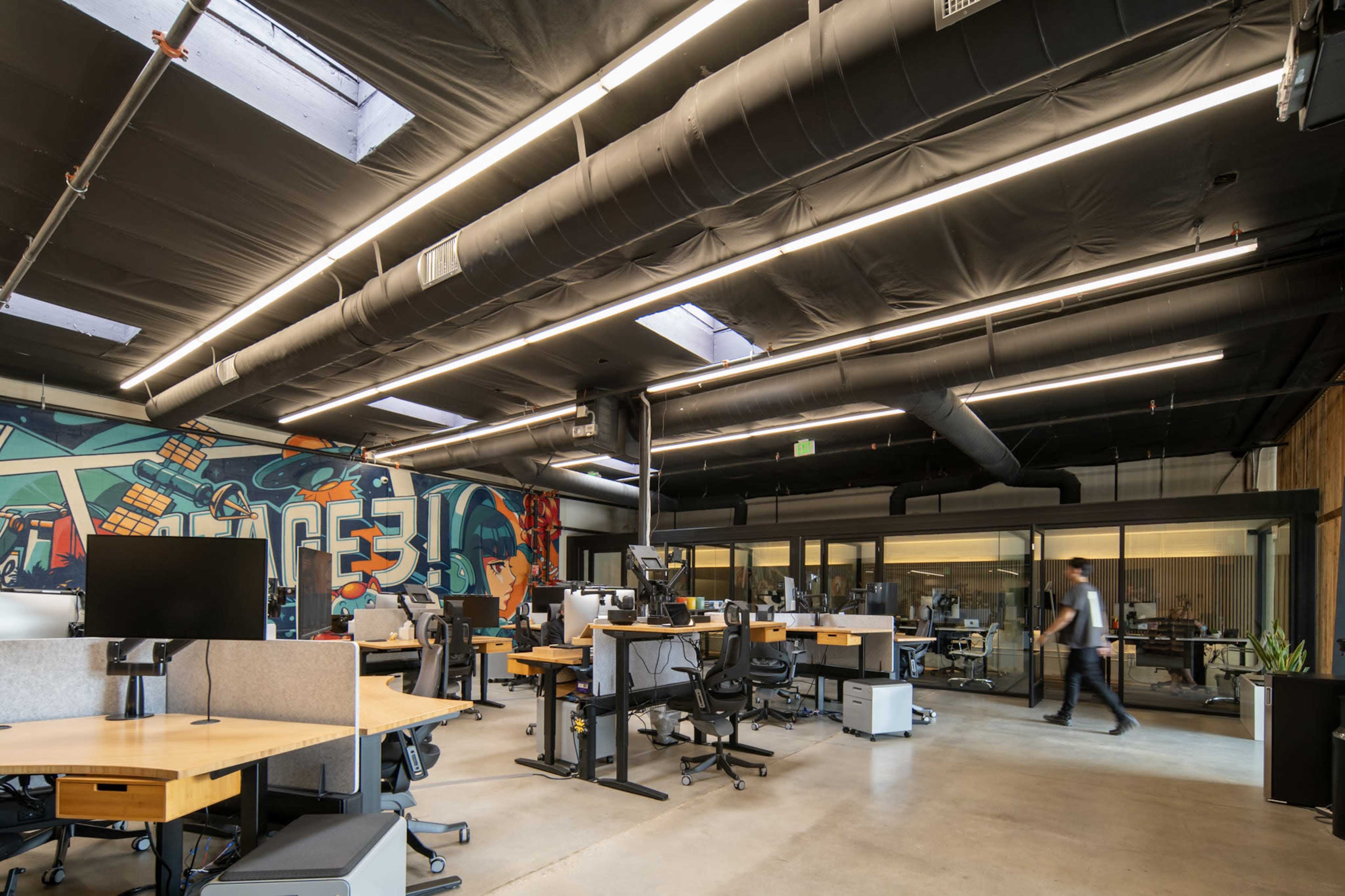 The image shows a modern office interior with multiple workstations, large windows, and a colorful mural on the wall.