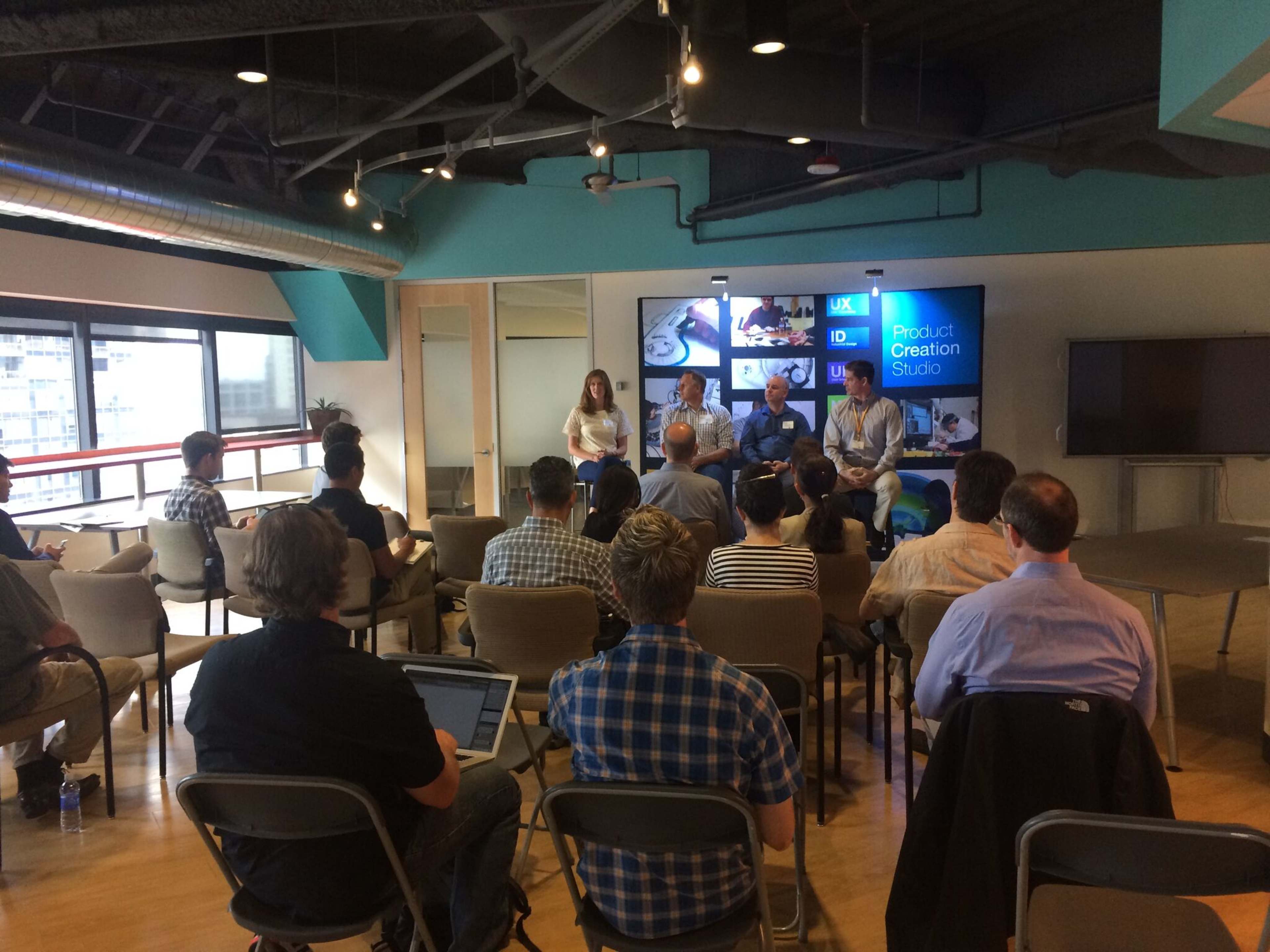 A panel discussion is taking place in a modern office space, with audience members seated in front of a group of speakers on a stage.