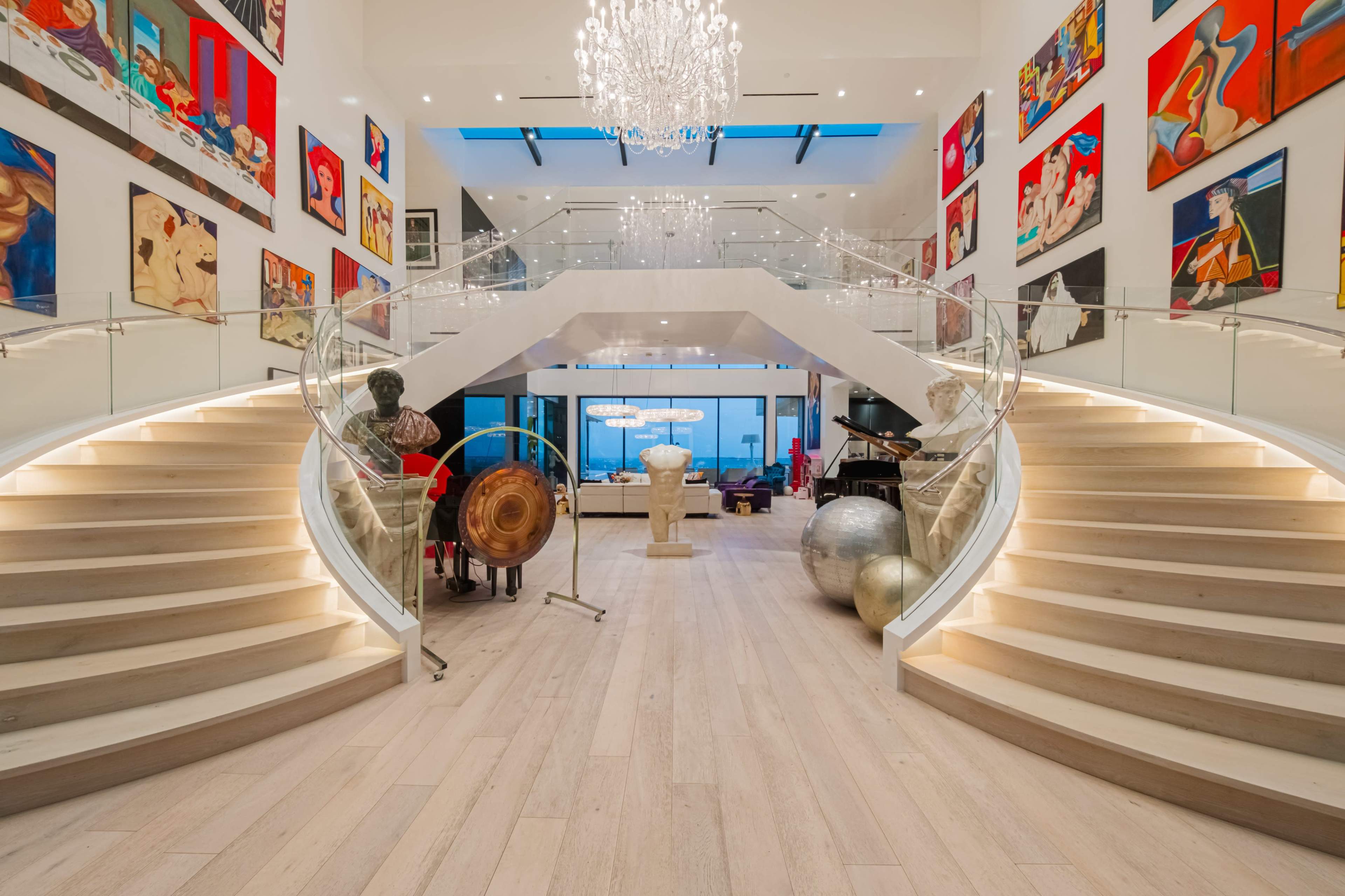 The image shows a spacious foyer with a sweeping double staircase, large artworks on the walls, and contemporary furnishings.