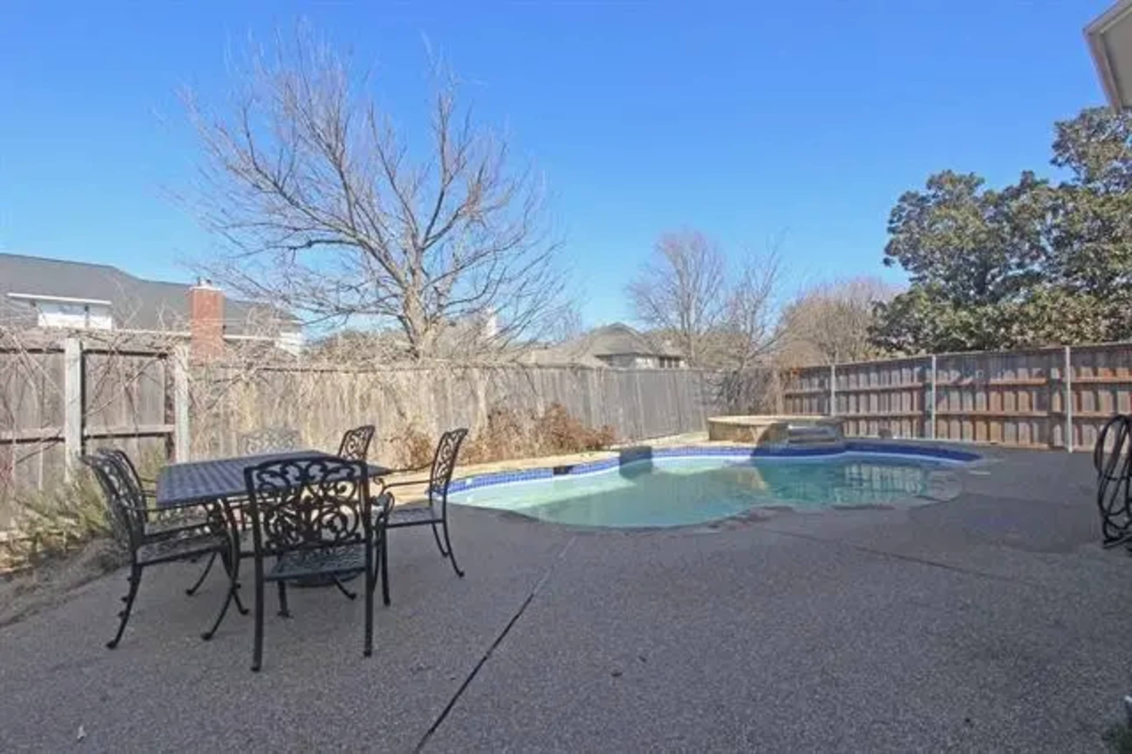 A backyard with a swimming pool surrounded by a concrete patio and outdoor furniture, enclosed by a wooden fence.
