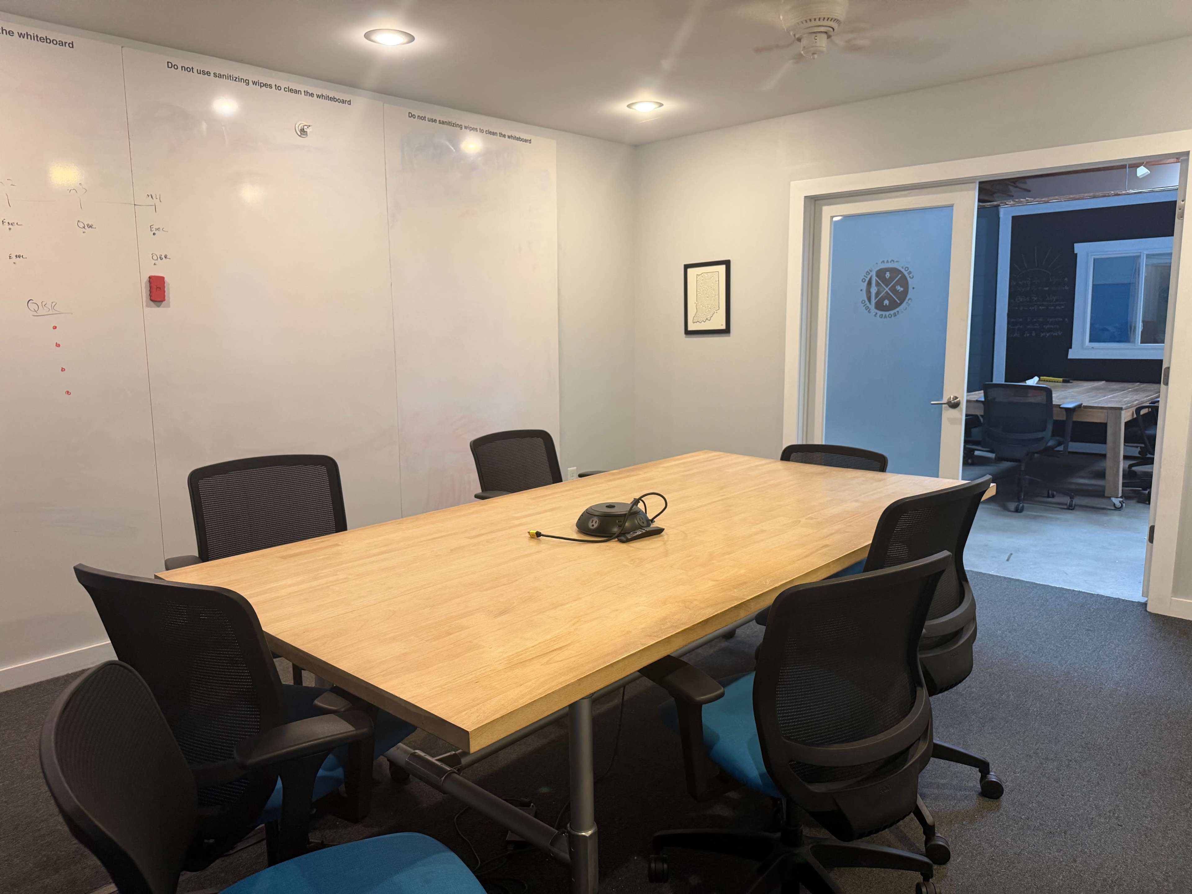 A conference room features a large wooden table surrounded by six black office chairs, with whiteboard walls and a door leading to another room.