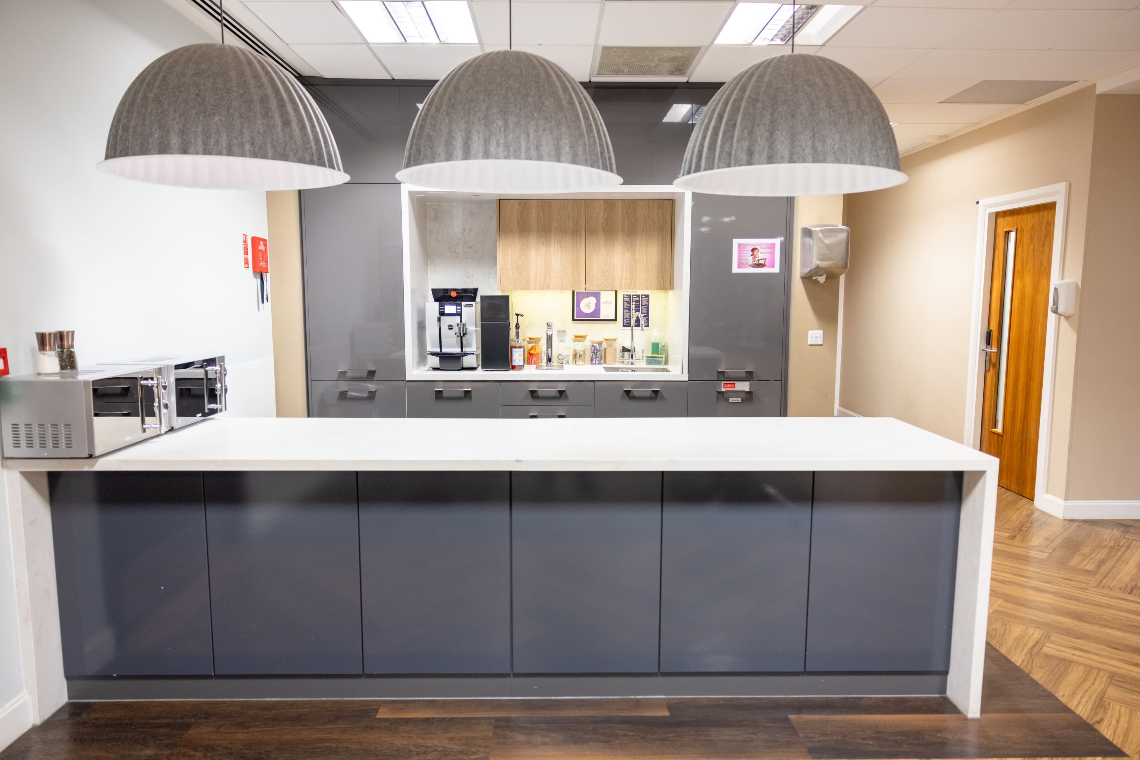 A modern kitchen area with a sleek gray countertop, three large pendant lights, and appliances including a coffee machine and microwaves.