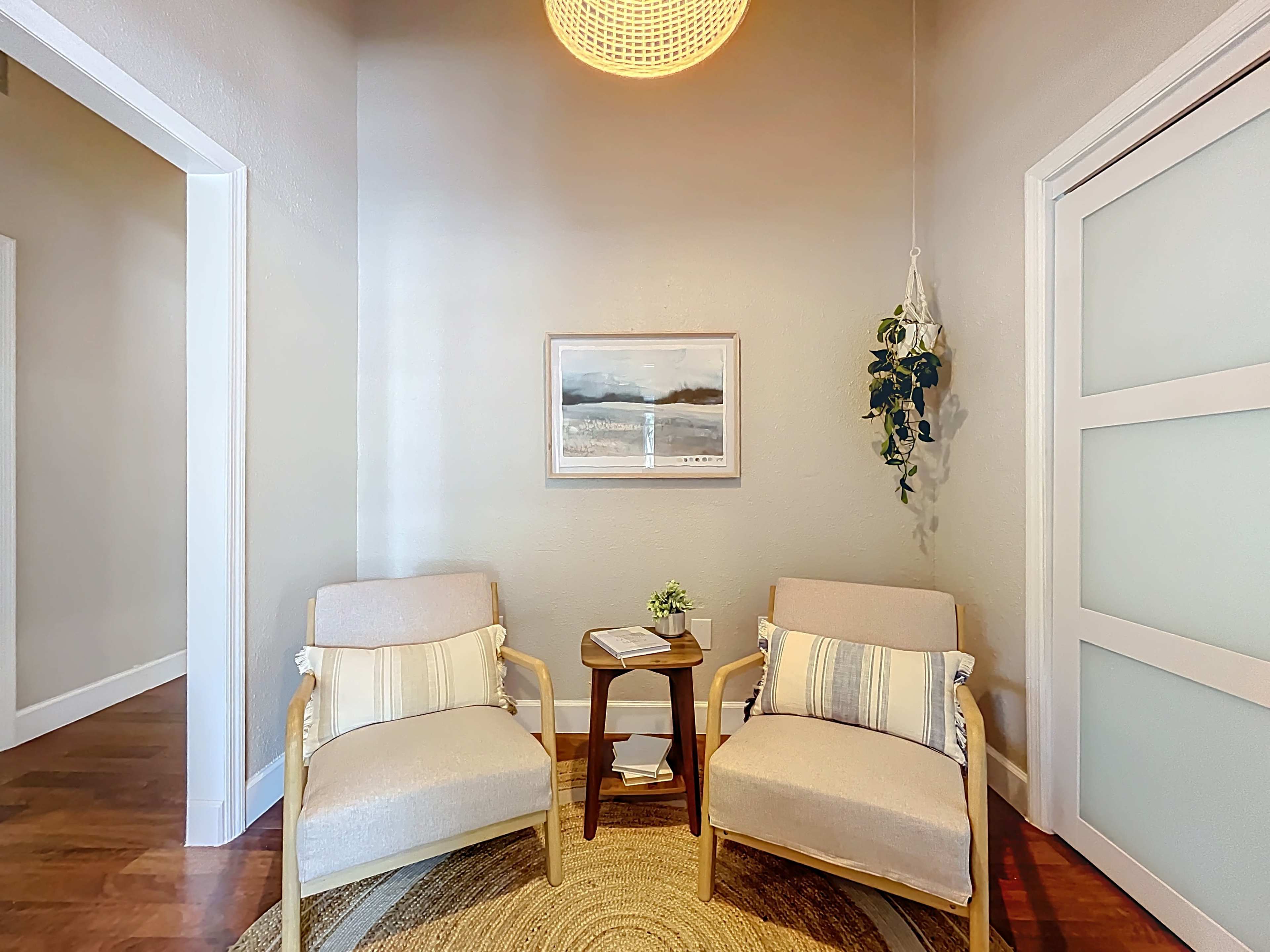 A cozy seating area features two beige chairs, a small round table, and a framed landscape picture on the wall, accompanied by a hanging plant.