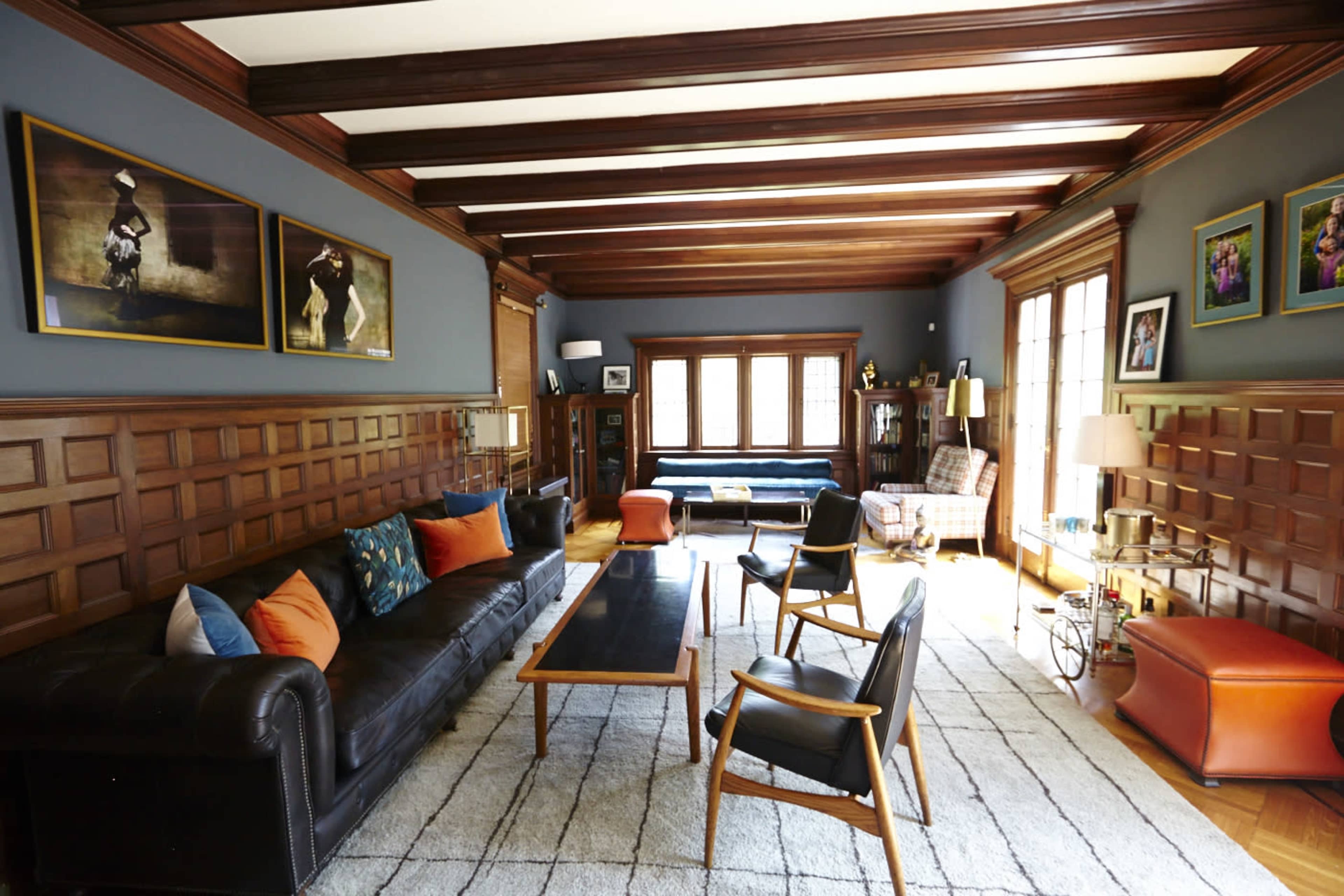 The living room features a dark leather sofa, wooden beams on the ceiling, a mix of chairs, and large windows allowing natural light.