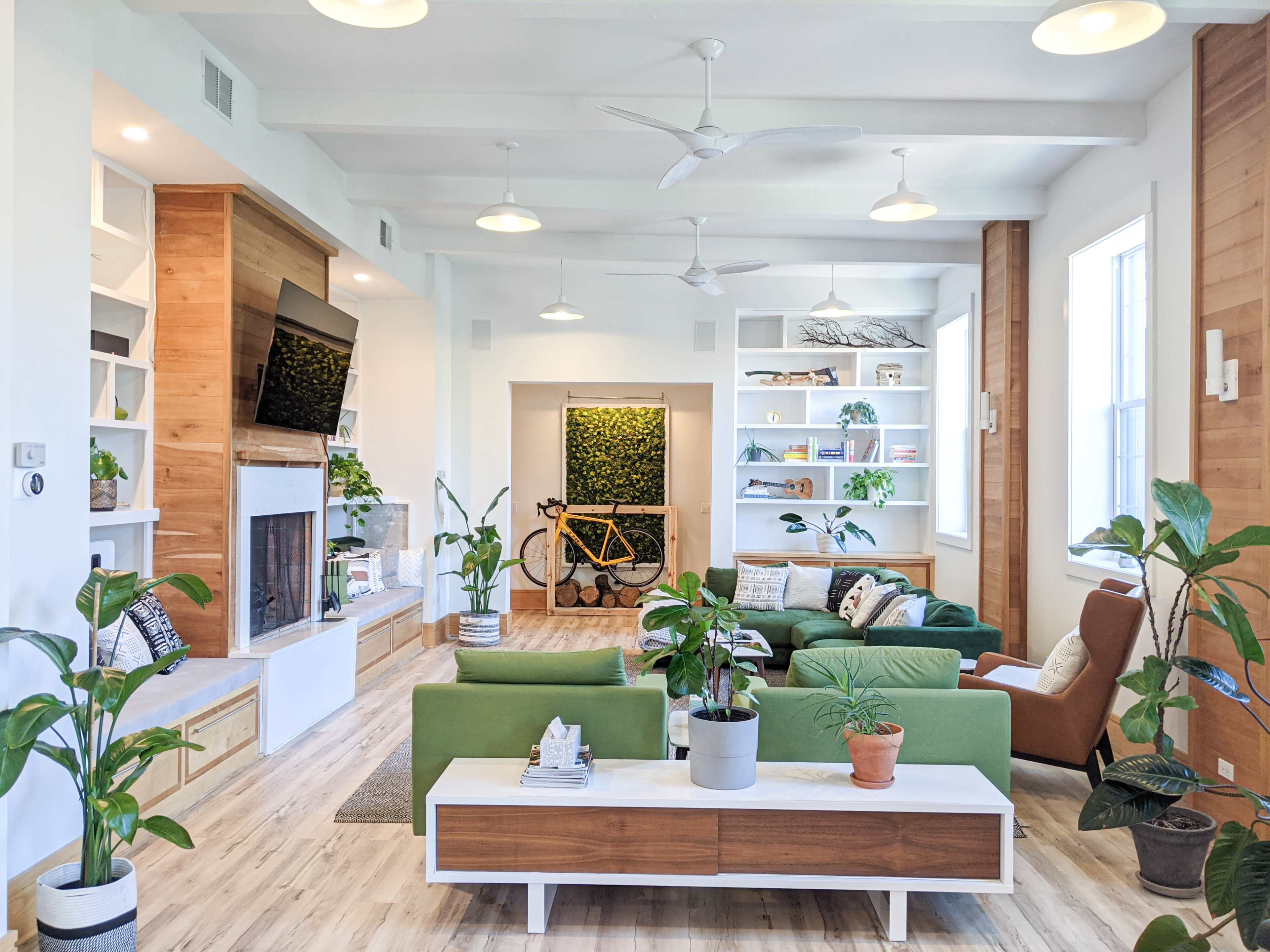 The living room features green sofas, a wooden coffee table, a fireplace, shelves filled with plants and decor, and a bicycle leaning against a wall.