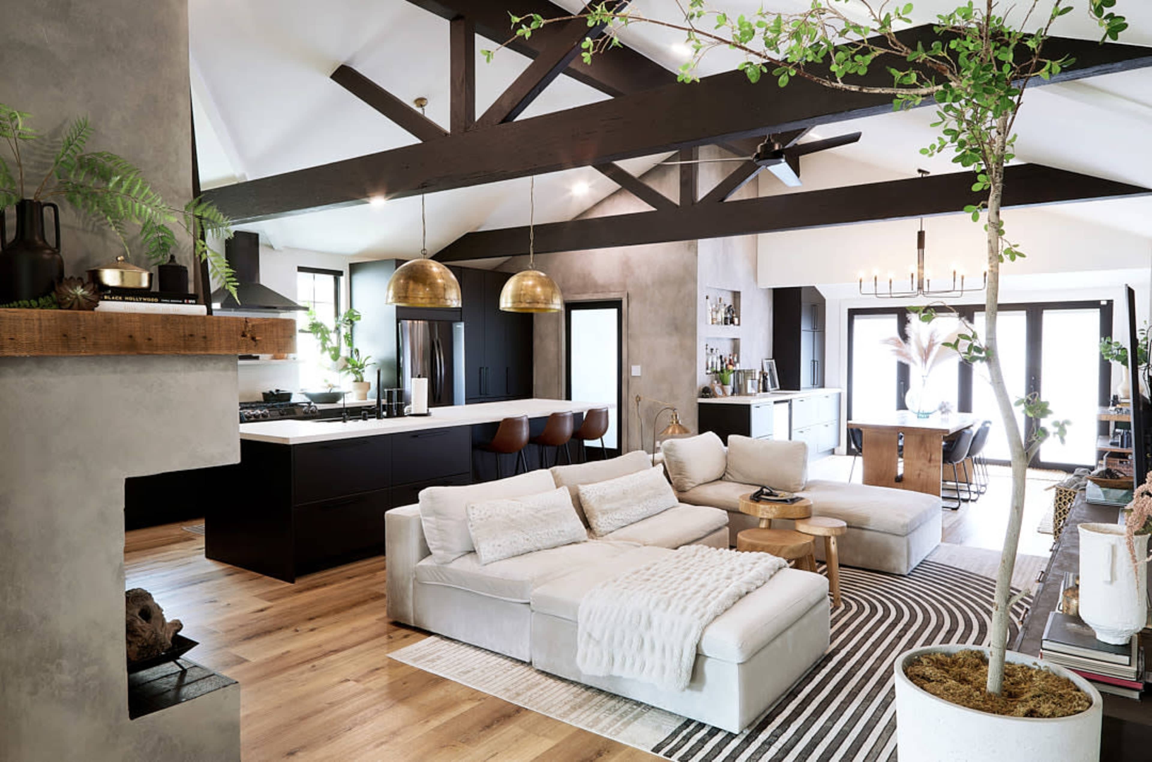 A modern open-concept living area features a large sectional sofa, wooden flooring, and a mix of dark and light cabinetry in the kitchen.
