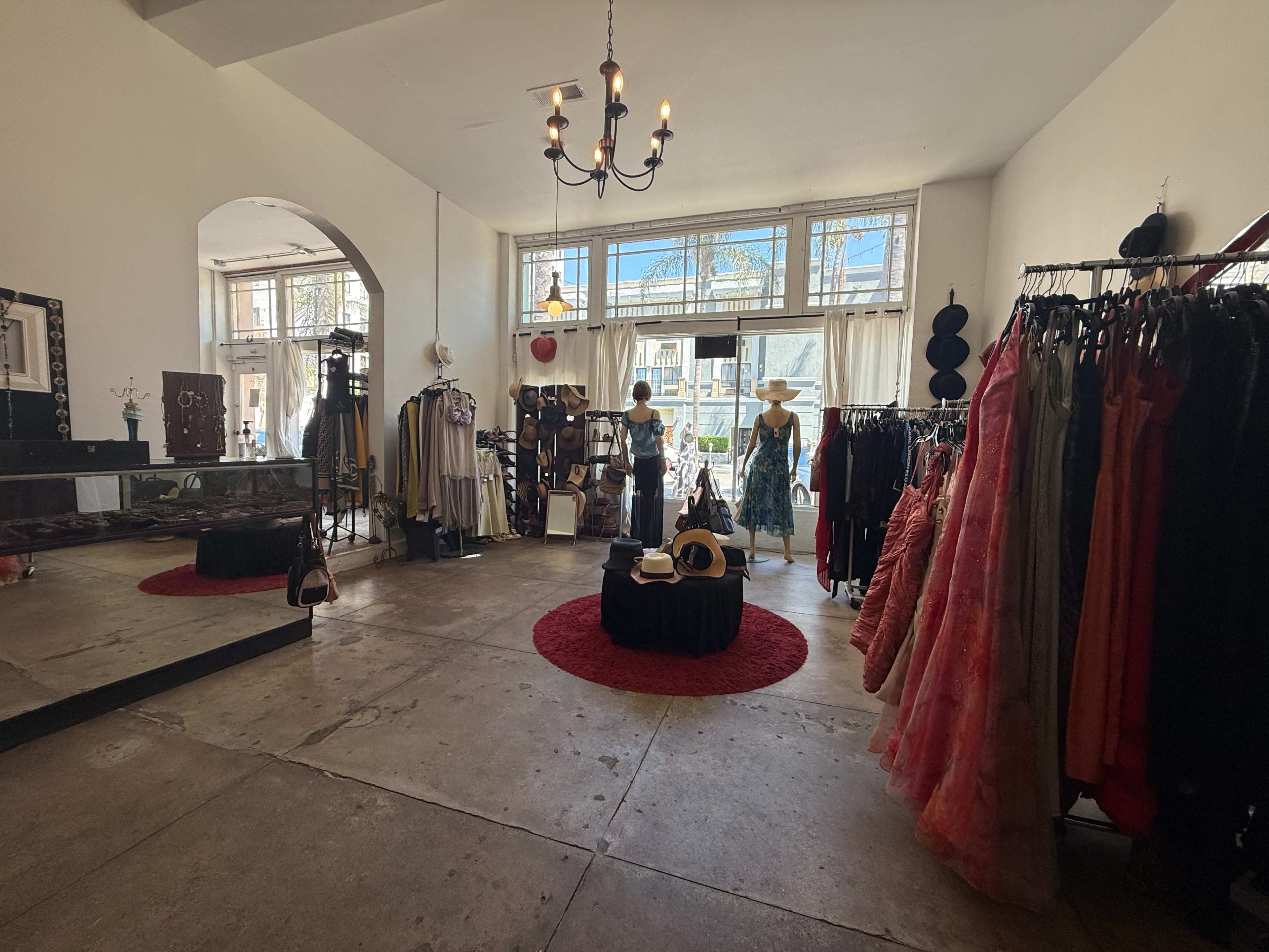 The interior of a boutique showcases clothing racks filled with various garments, a round display with hats, and a bright window that allows natural light to fill the space.
