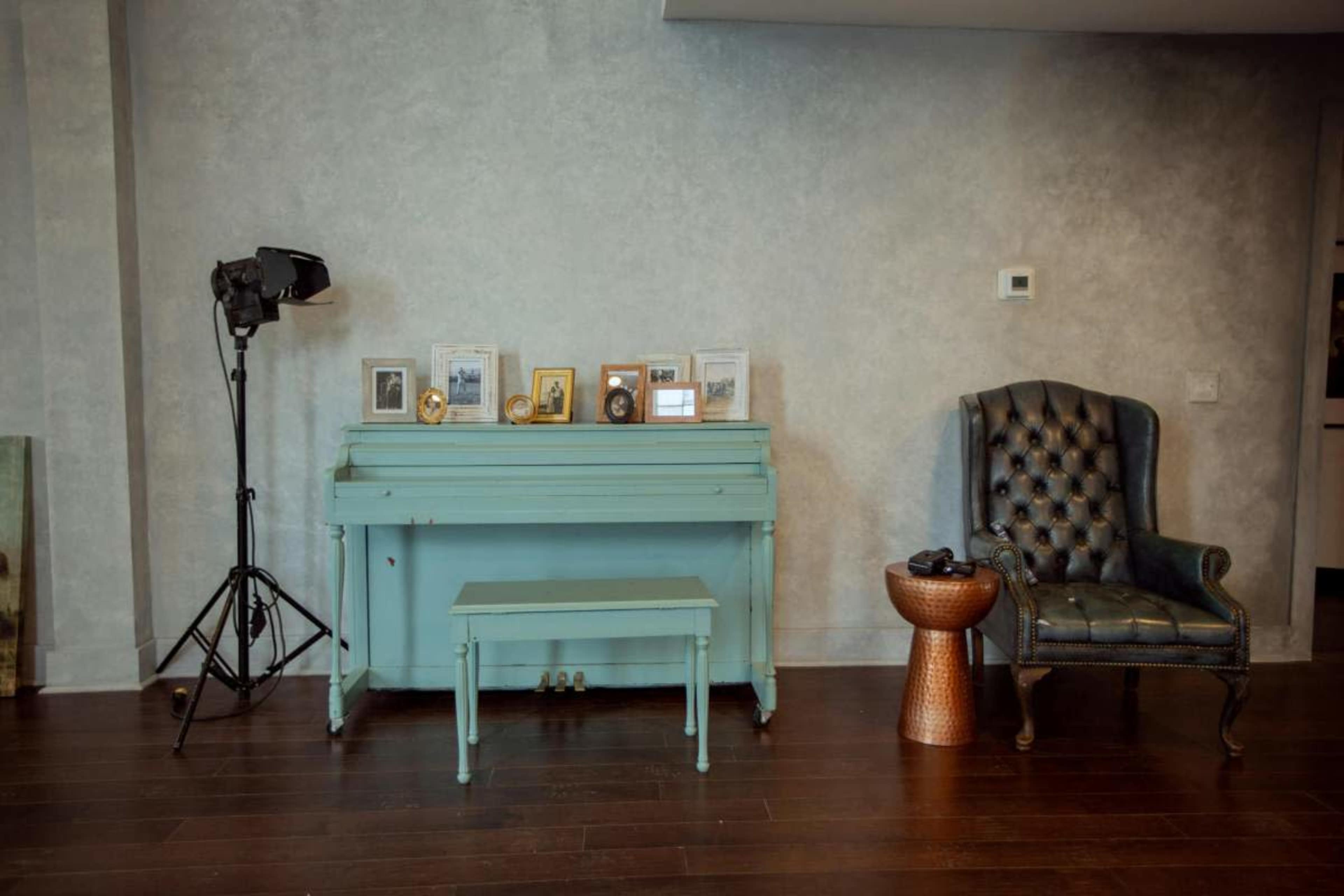 A light blue piano is positioned next to a wooden side table and a tufted dark chair in a room with gray walls and hardwood flooring.