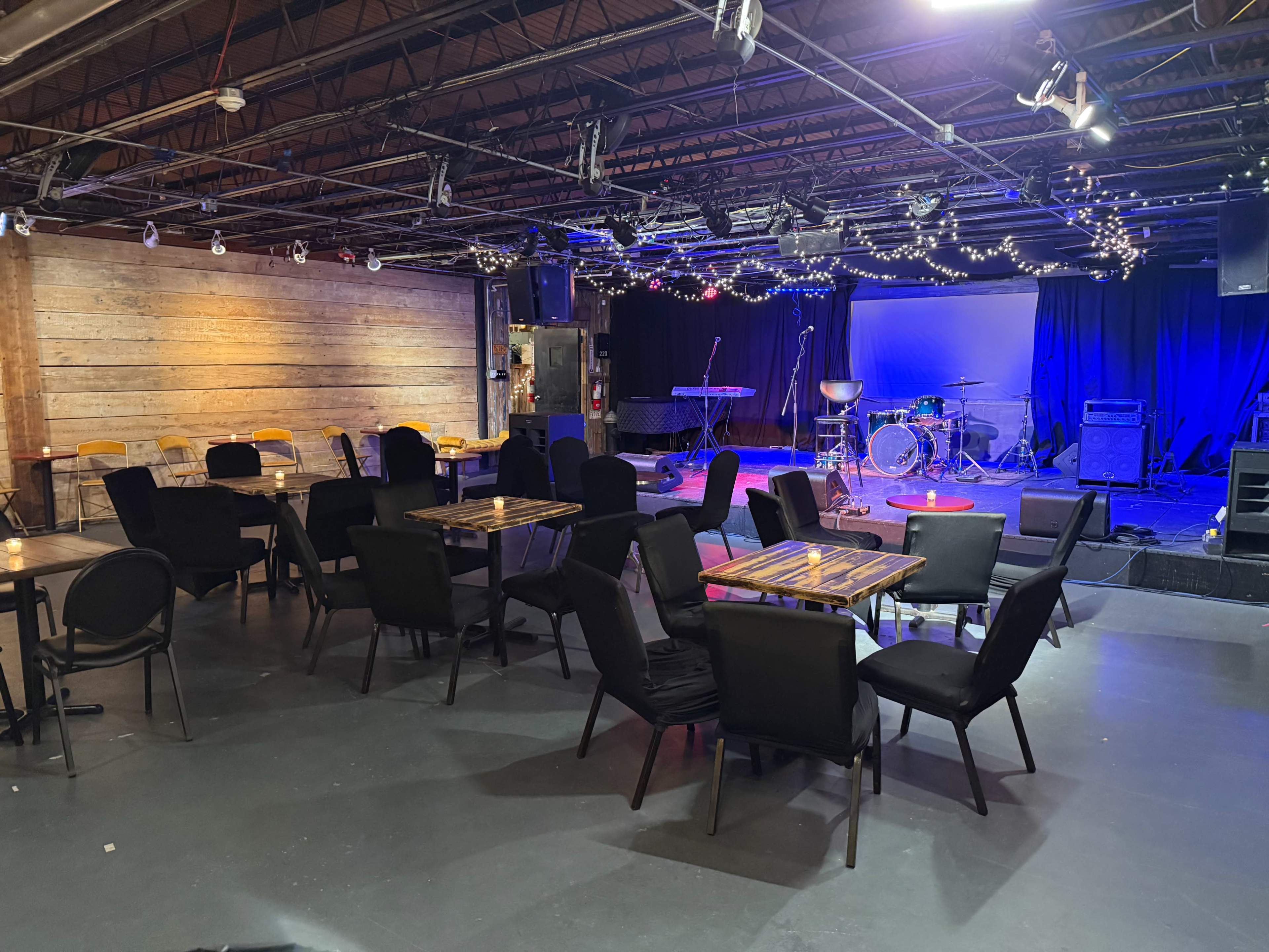 The image shows an empty music venue with tables and chairs arranged in front of a stage equipped with instruments and vibrant lighting.