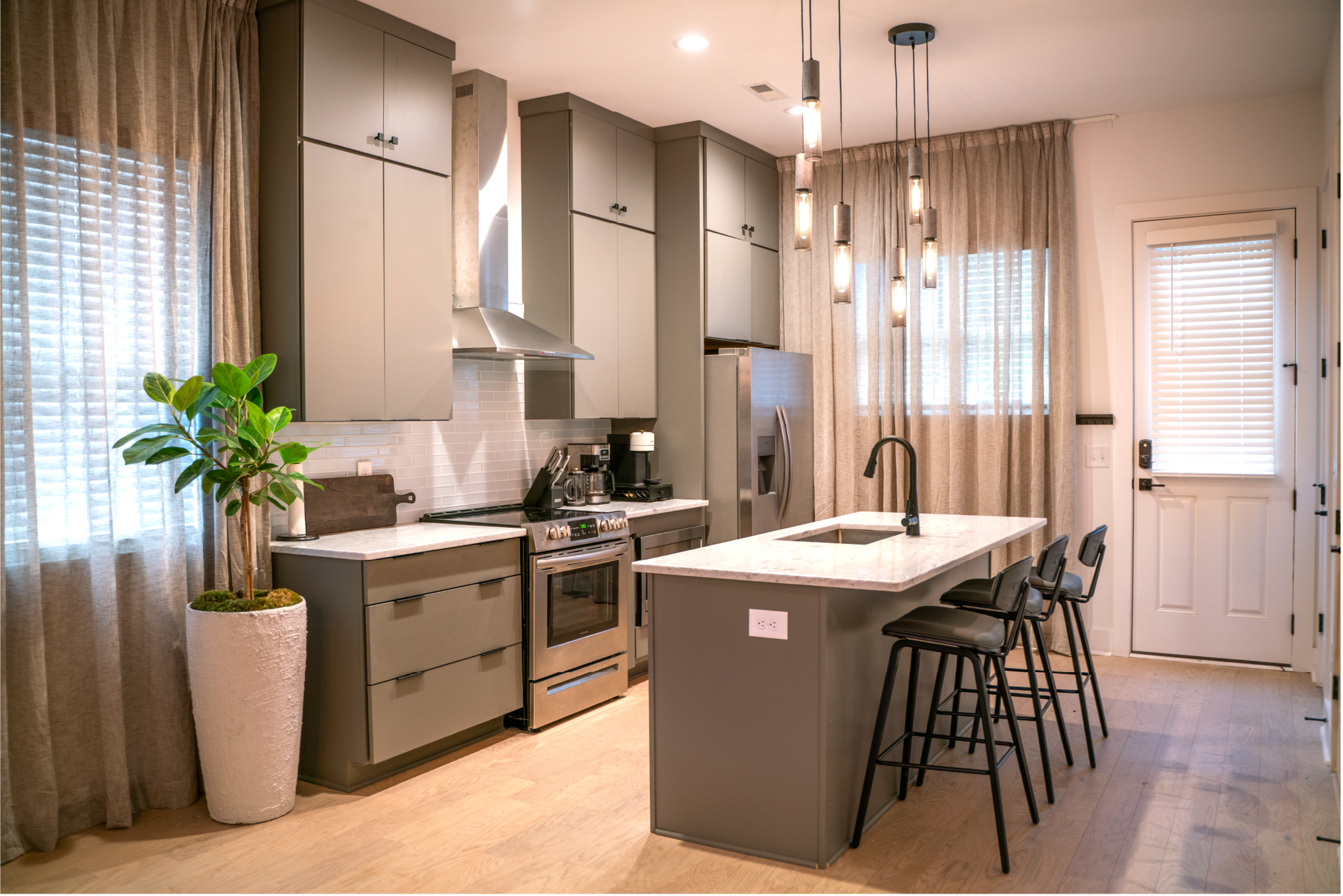 A modern kitchen features gray cabinetry, stainless steel appliances, and a central island with bar stools, illuminated by pendant lights.