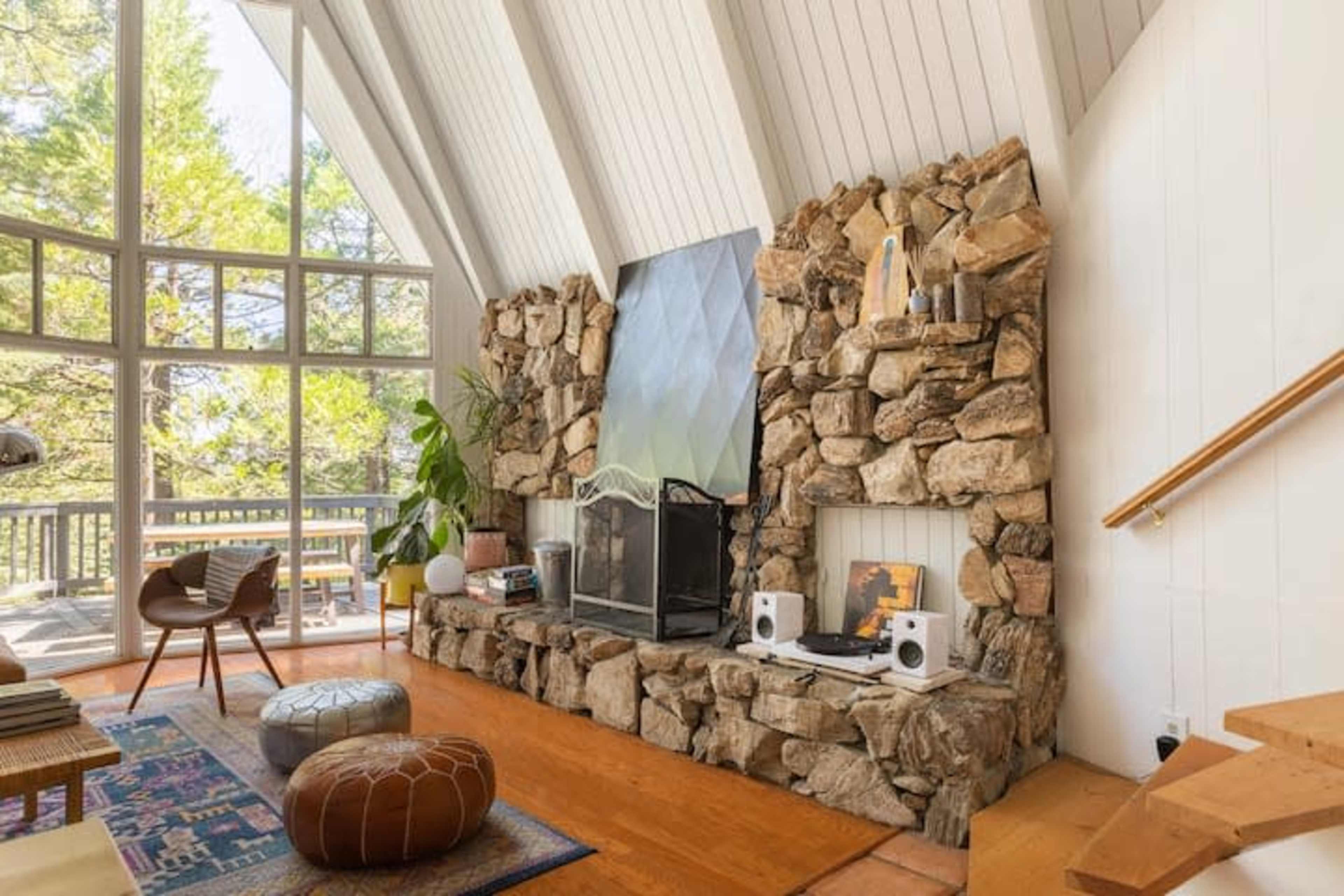 A modern living room features a stone fireplace with a large artwork above it, surrounded by large windows and wooden flooring.