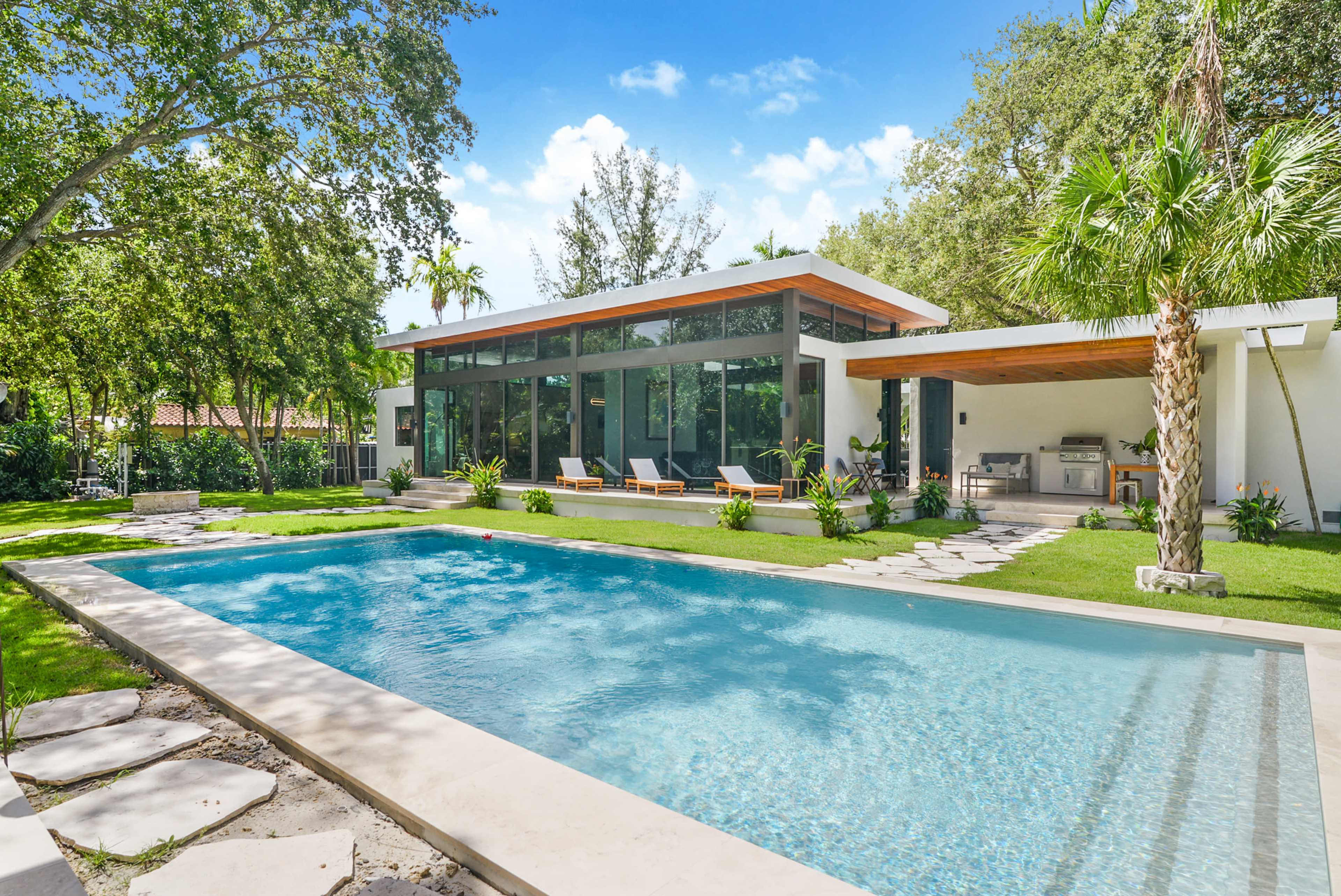 A modern house with large glass windows overlooks a swimming pool surrounded by landscaped greenery and patio chairs.