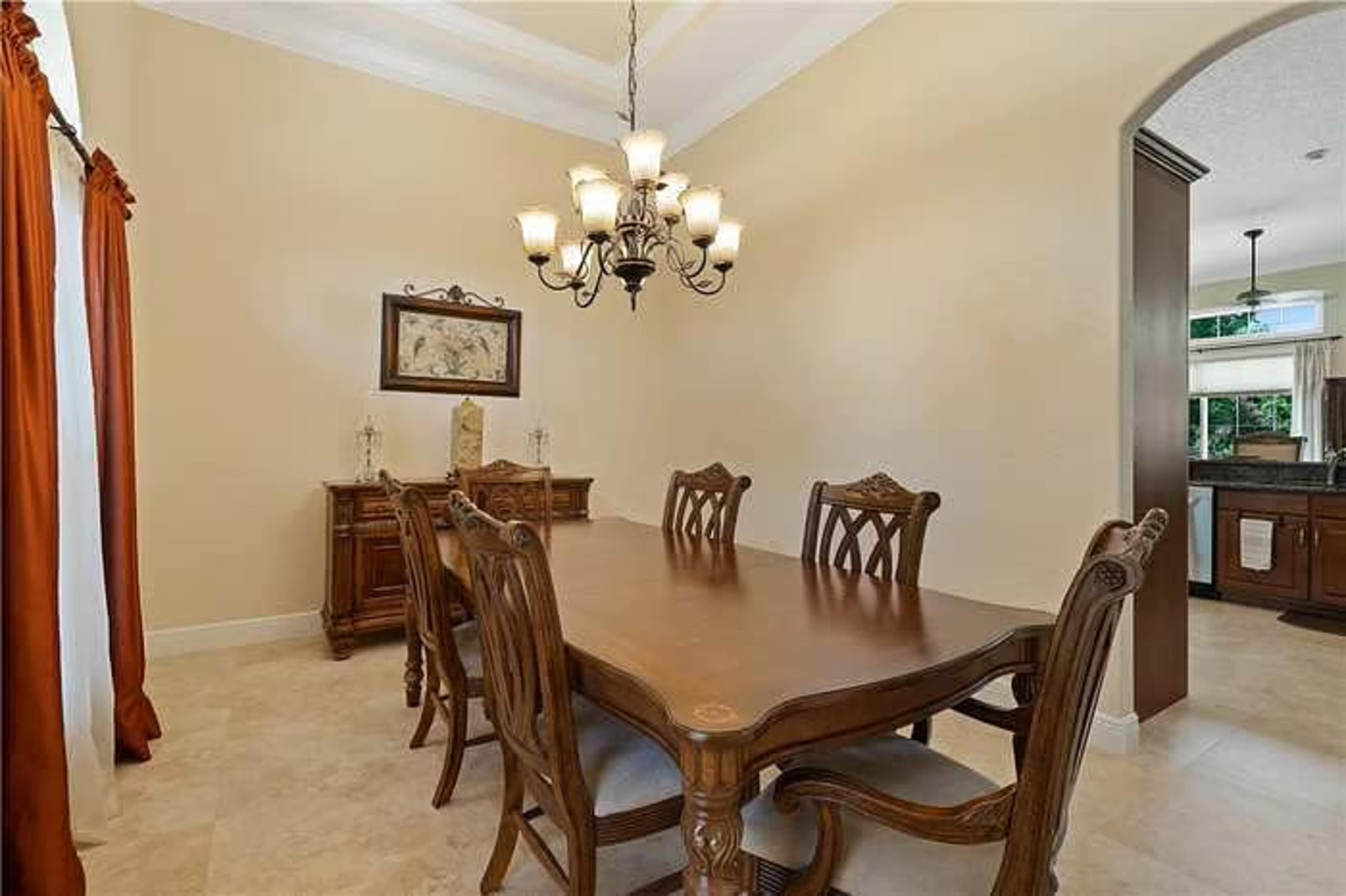 A dining room features a large wooden table surrounded by elegant chairs, illuminated by a chandelier, with a sideboard and windows dressed in curtains.