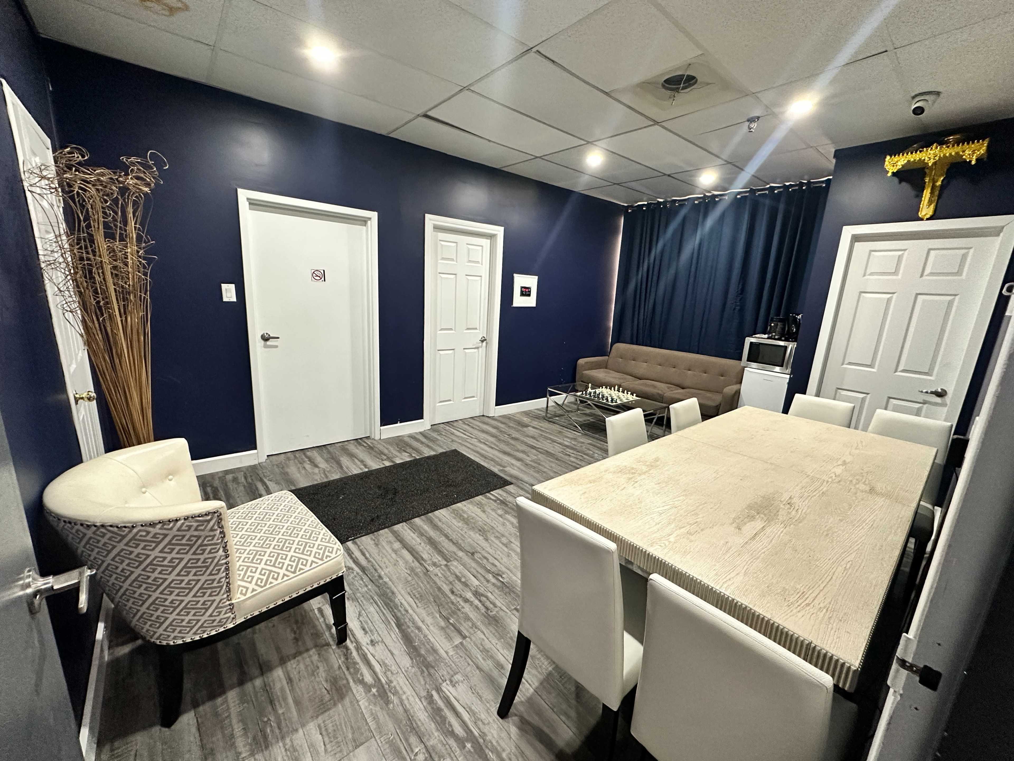 The room features a brown sofa, a white dining table surrounded by chairs, and multiple doors against dark blue walls.