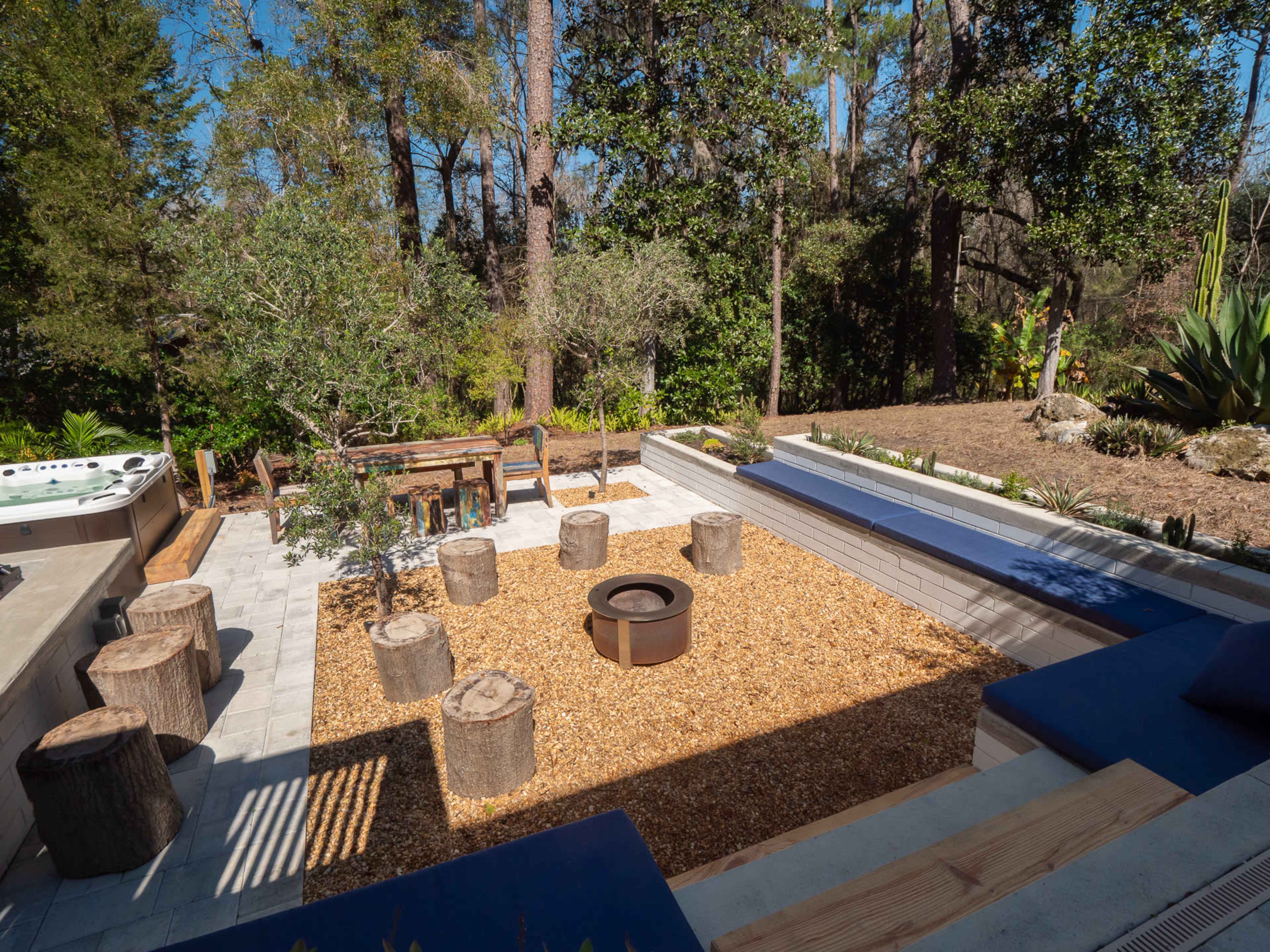 The image shows a landscaped outdoor area with a fire pit surrounded by log seating, benches, and a dining table set among trees and greenery.