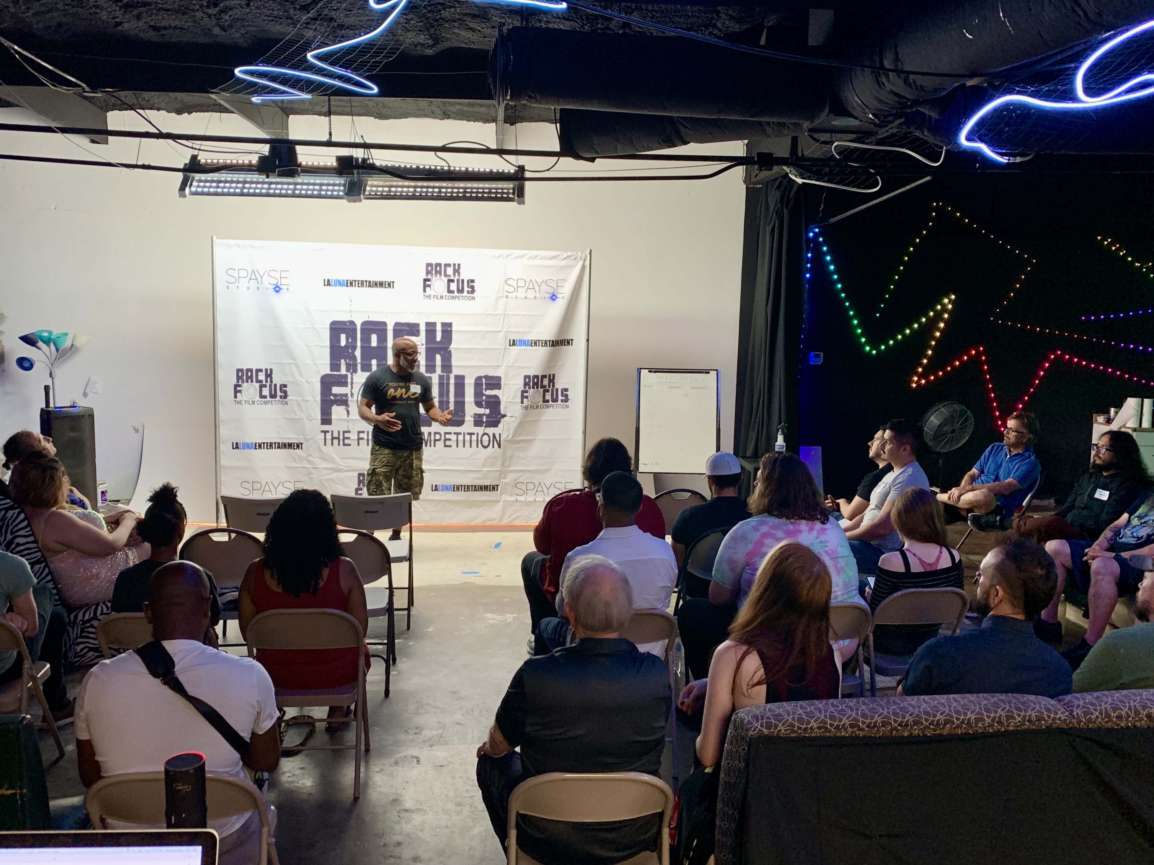 A speaker stands on a stage in front of an audience at a film competition event in a dimly lit room.