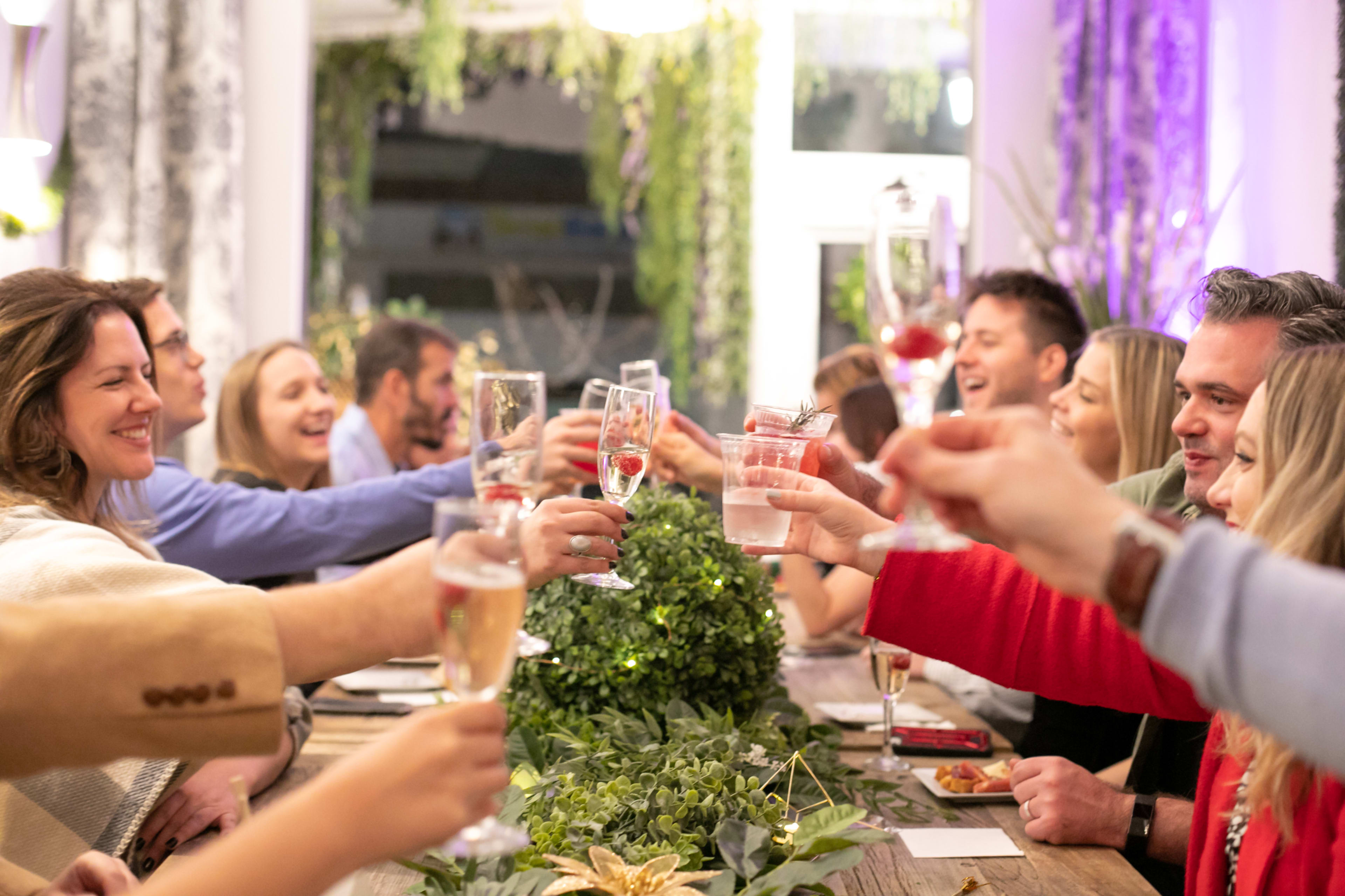 A group of people at a festive dinner is raising glasses in a toast around a long table decorated with greenery.