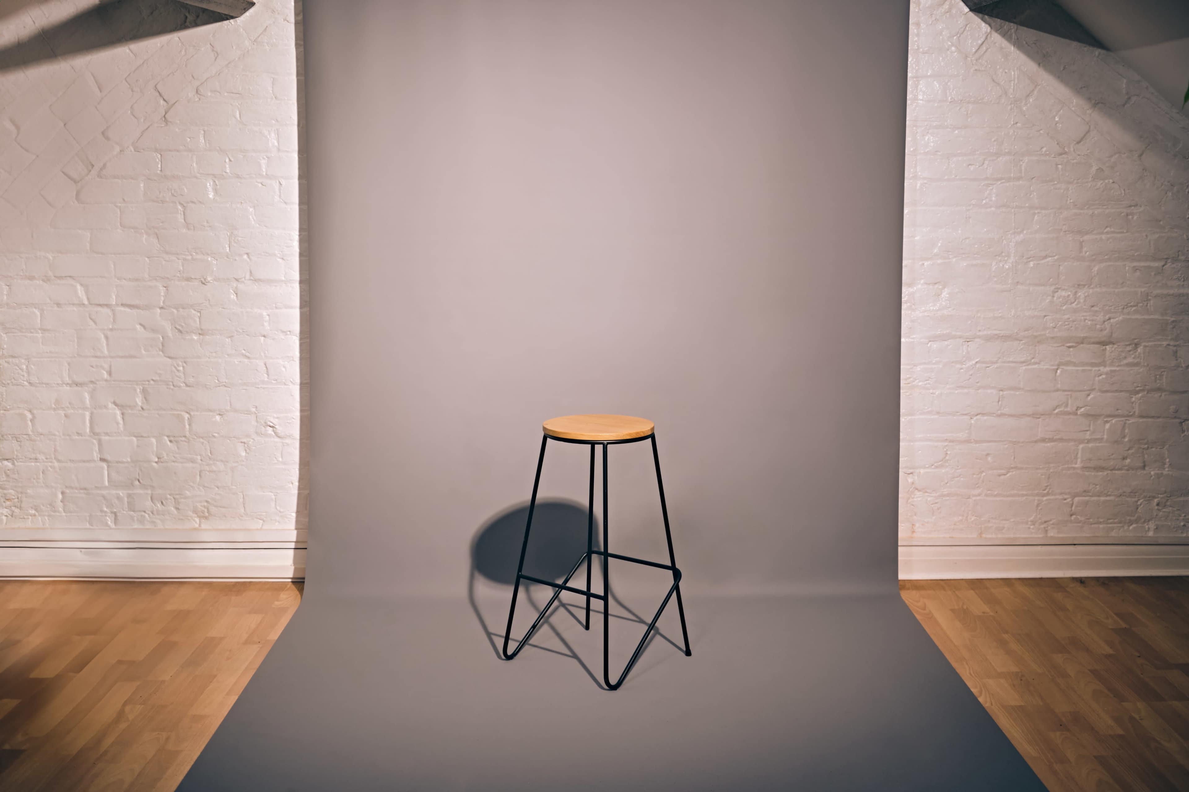 A wooden stool sits on a gray backdrop in a well-lit room with white brick walls.