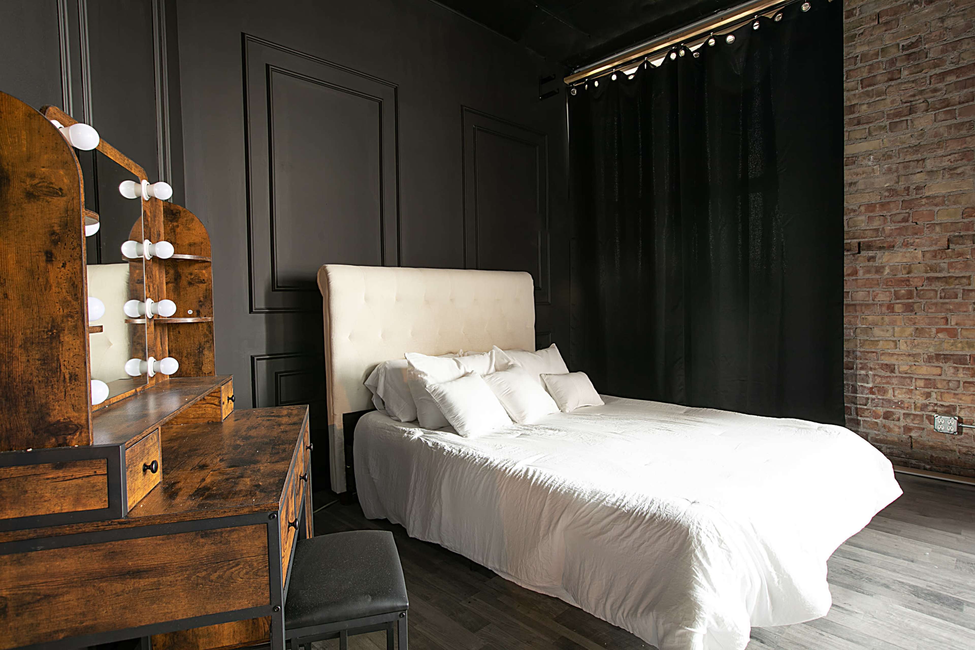 A minimalistic bedroom featuring a white bed with multiple pillows, a wooden dresser with lights, and a dark brick wall.