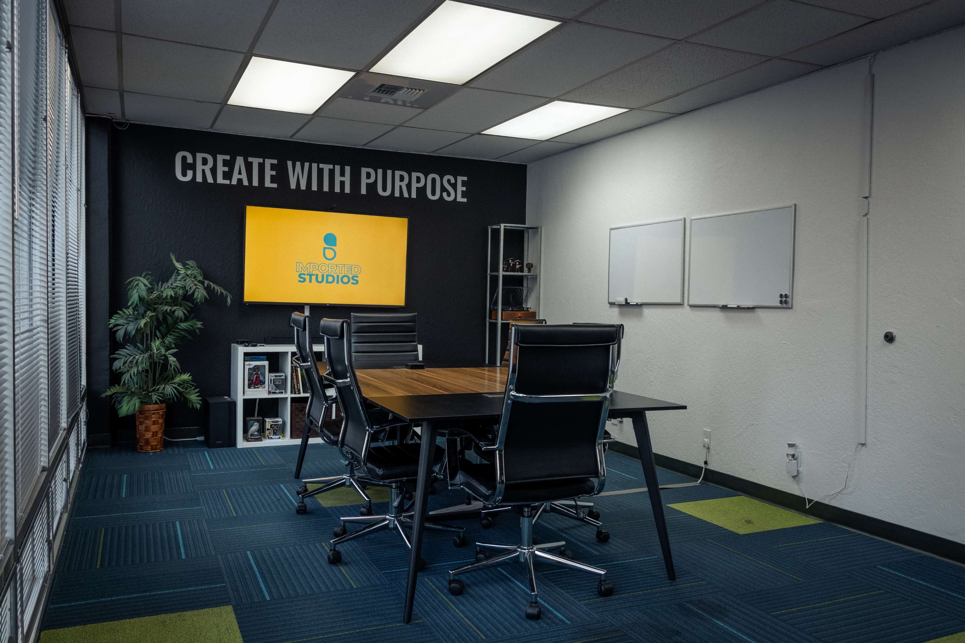 A conference room features a large rectangular table surrounded by modern chairs, with a wall-mounted screen displaying "CREATE WITH PURPOSE" and a decorative plant in the corner.