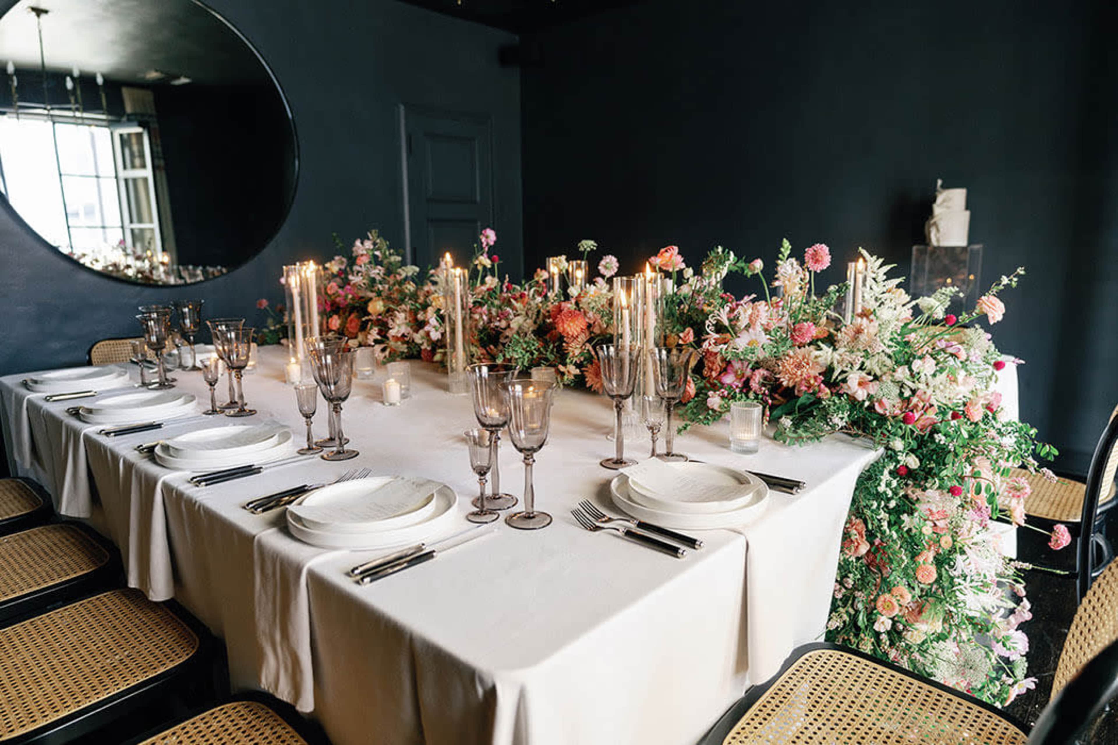 A long dining table is set with elegant tableware and decorated with a floral arrangement and candles on one side.