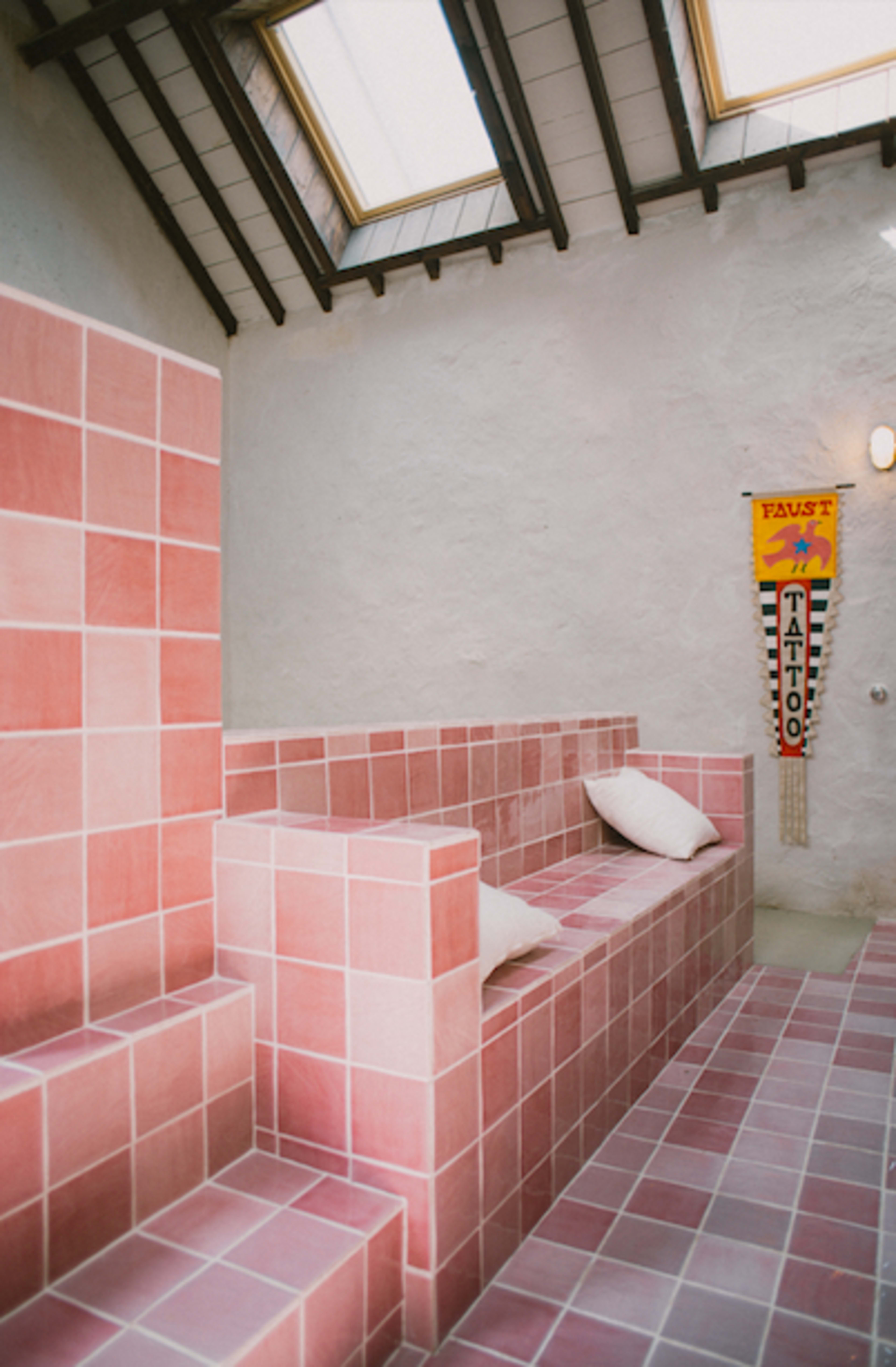 A room with pink tiled benches, white pillows, and a skylight, featuring a banner on the wall that reads "Faust Tattoo."