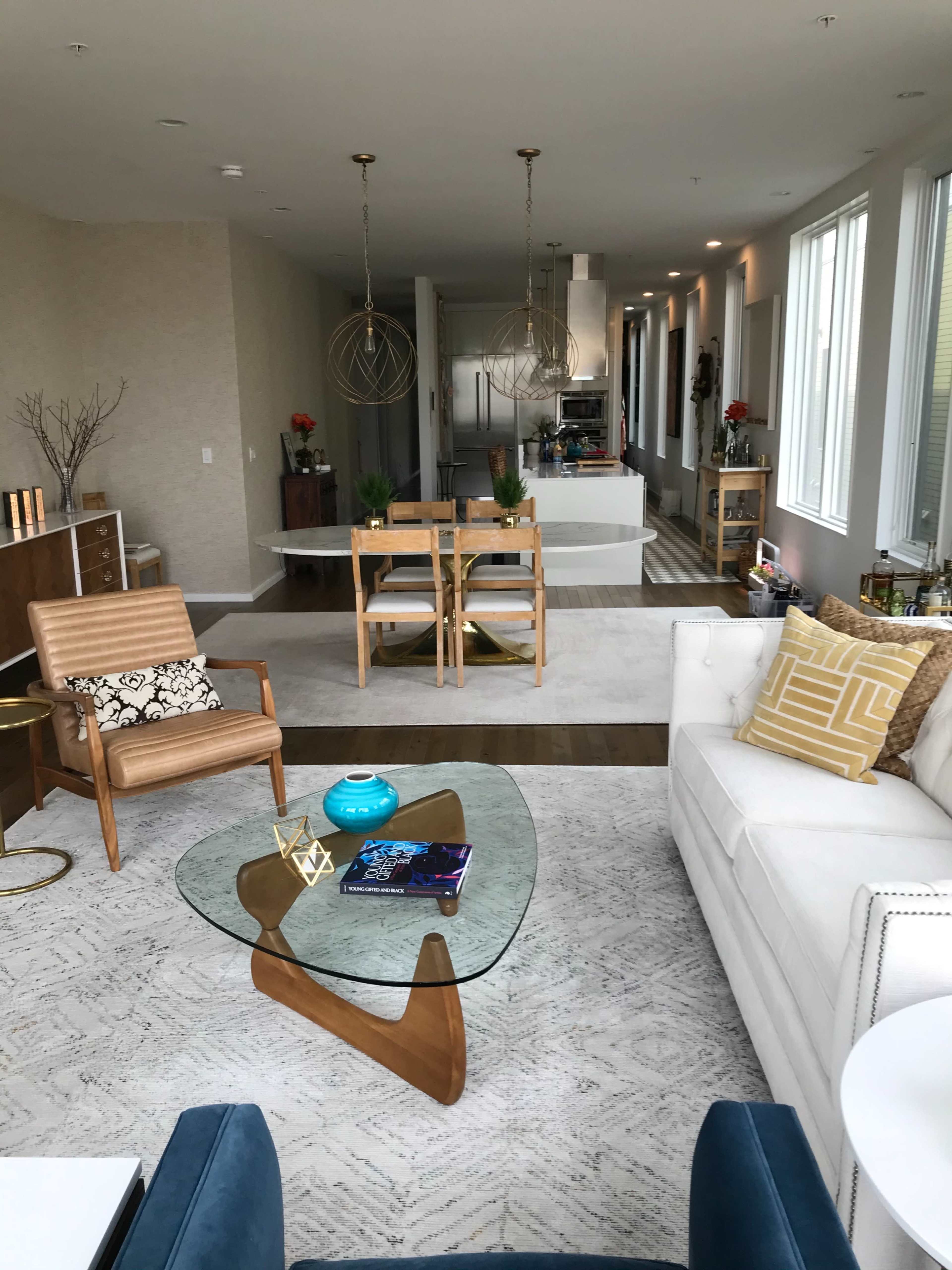 The image shows a modern living room with a glass coffee table, an armchair, and a white sofa, overlooking an open kitchen and dining area.