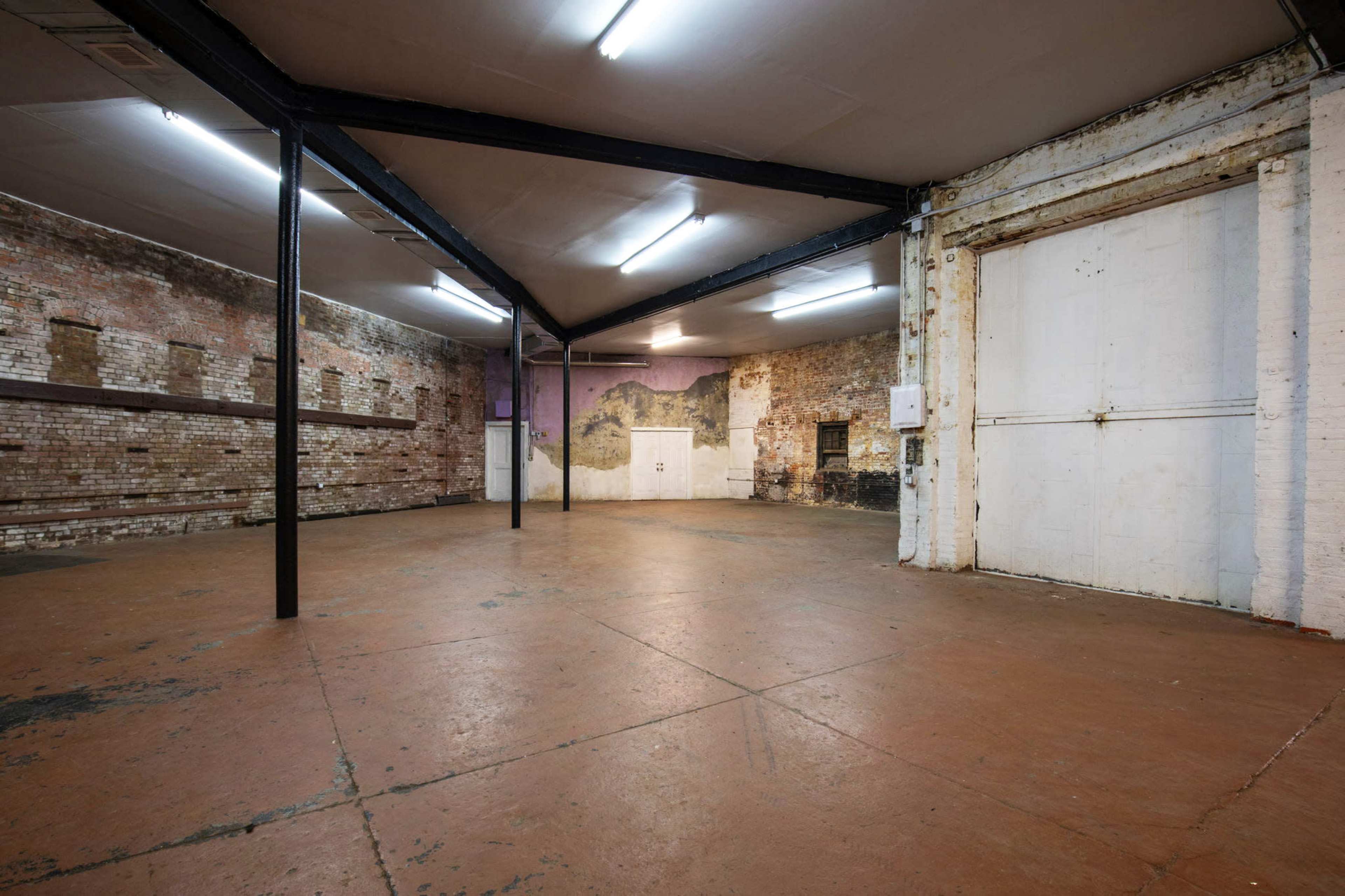 The image shows a spacious, empty industrial room with exposed brick walls and concrete flooring, featuring overhead lighting and a large entryway.
