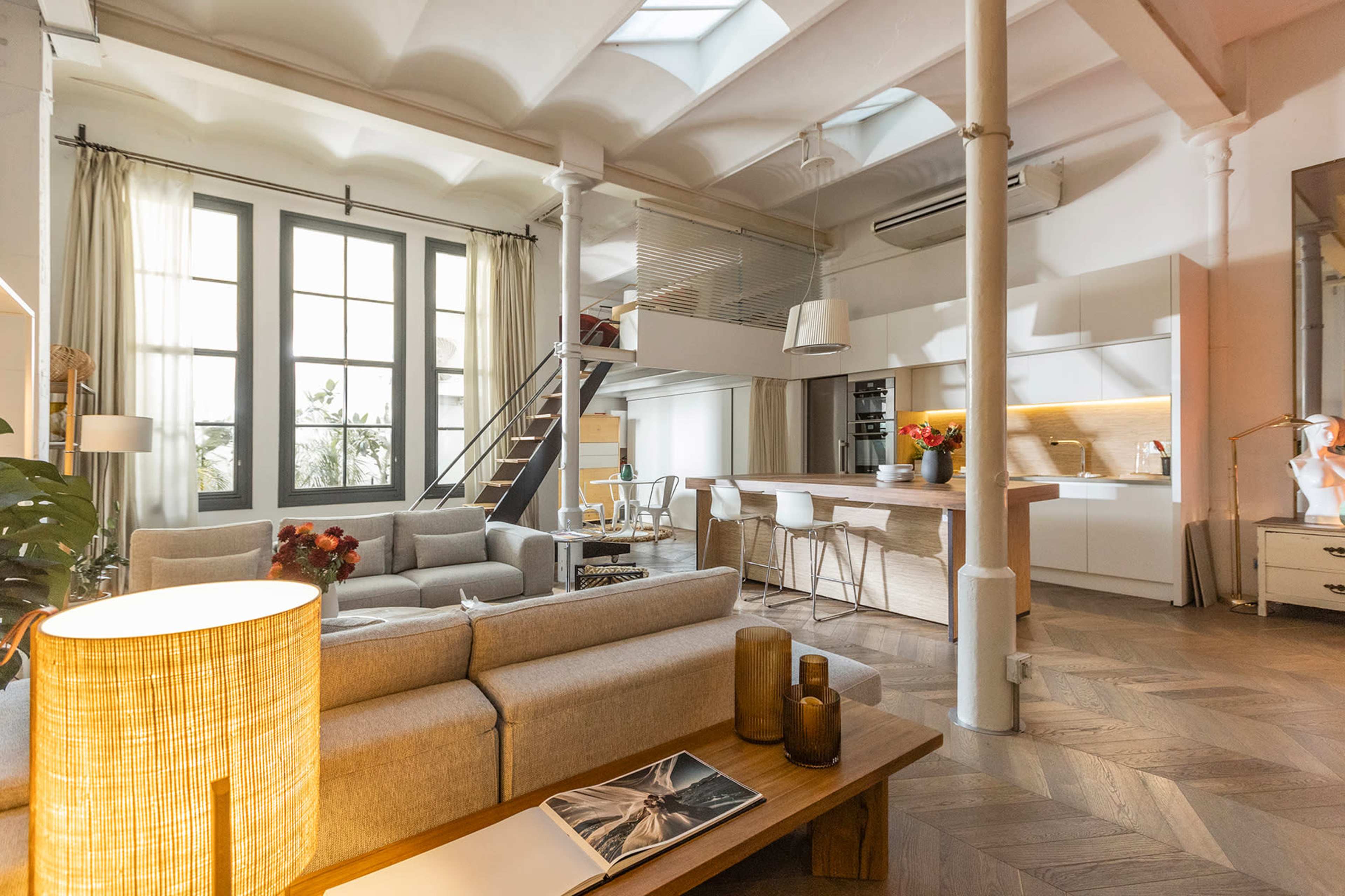 The image shows a spacious, modern living room with large windows, an open kitchen area, and a staircase leading to a loft.