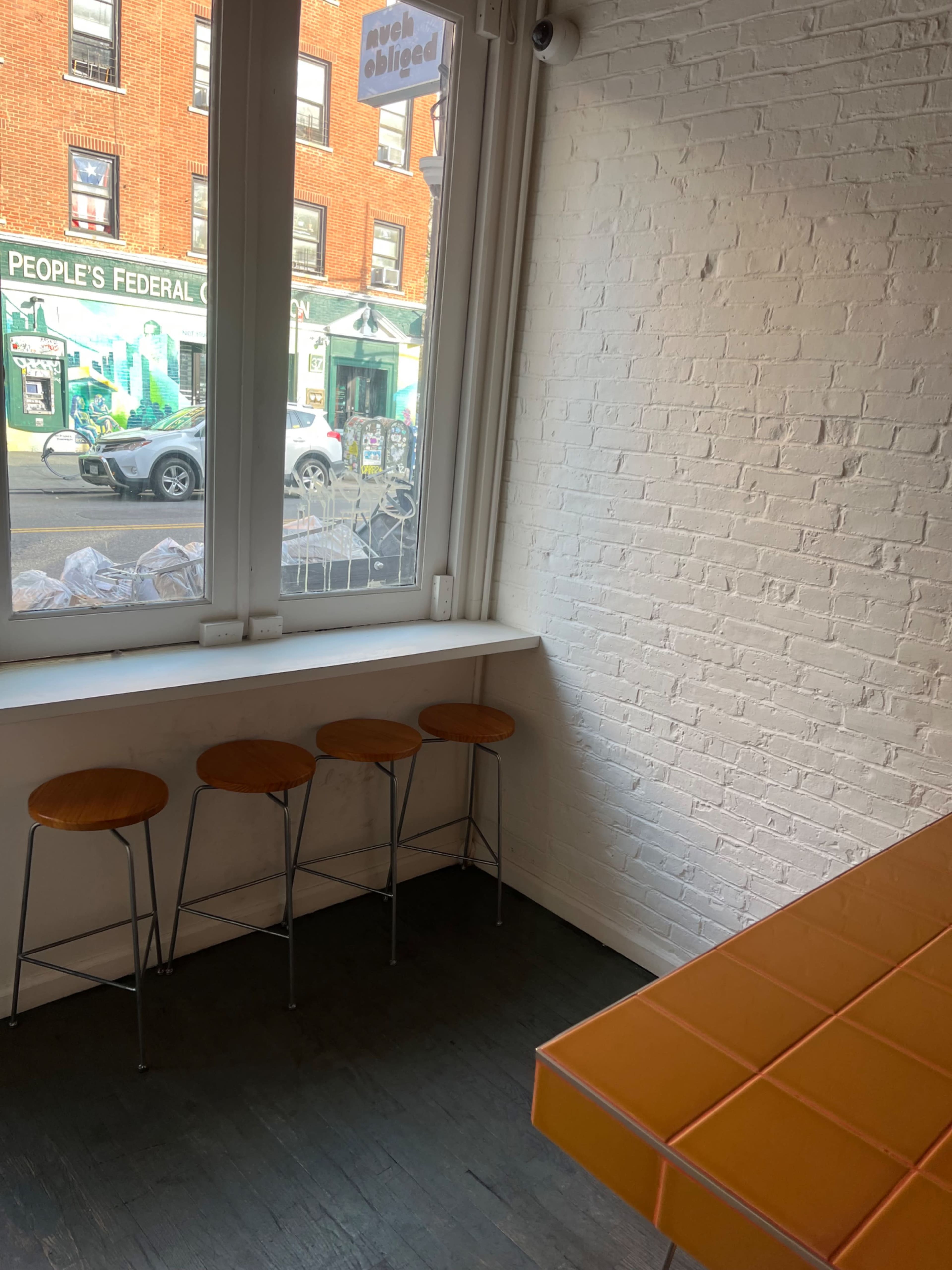 The image shows a bright, minimalist interior with three wooden stools lined up against a white-brick wall and a window overlooking a street with buildings.