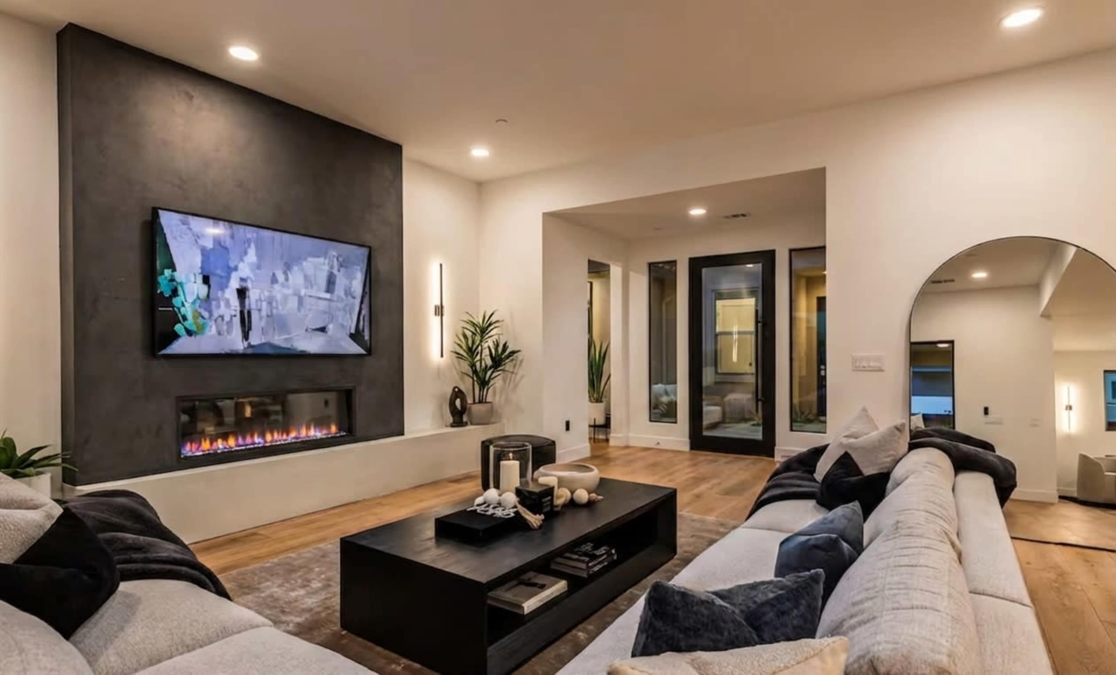 A modern living room features a large flat-screen TV mounted on the wall above a sleek fireplace, with a coffee table in the center and comfortable sofas arranged around it.
