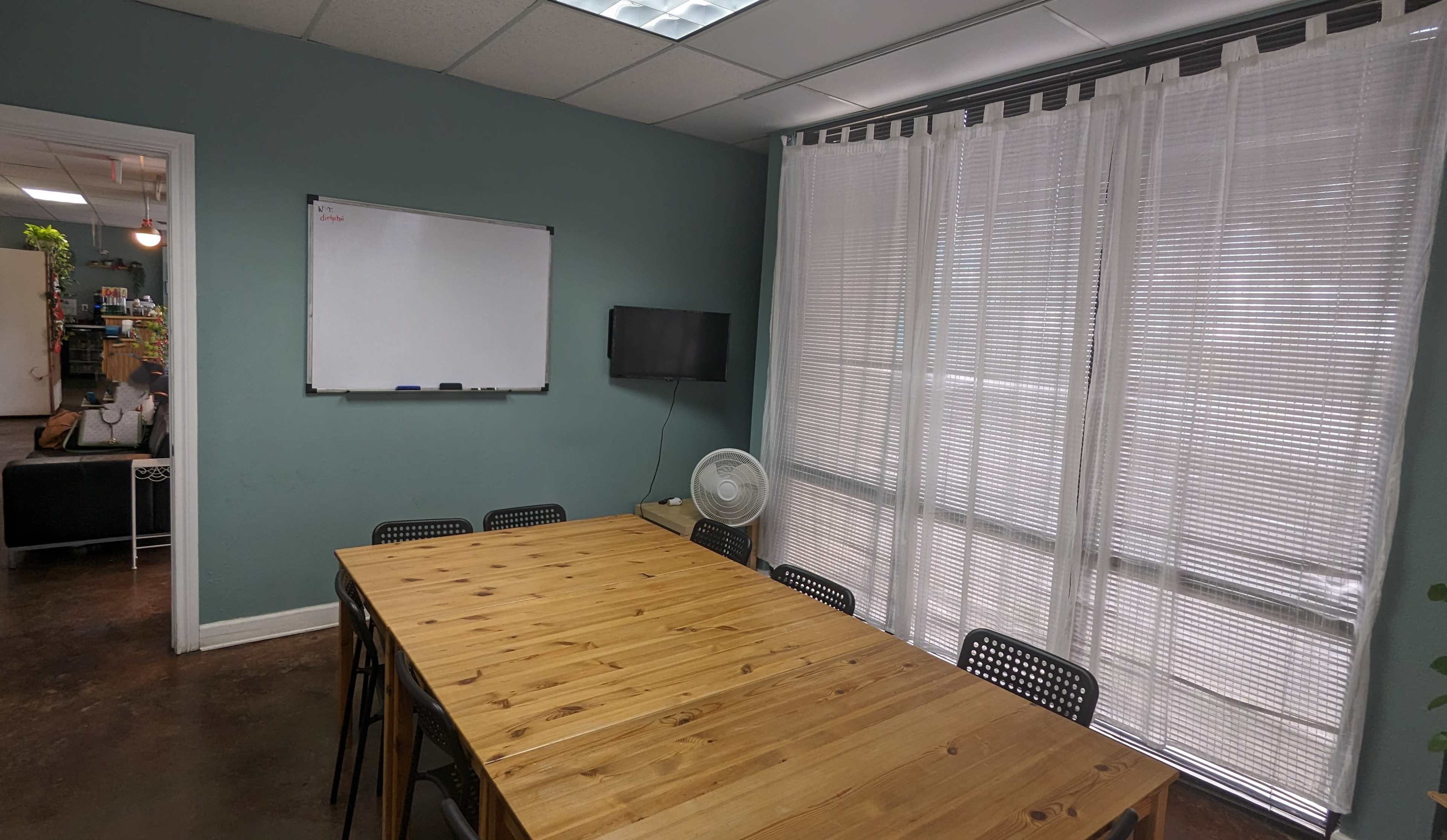 The image shows a meeting room with a wooden table, black chairs, a whiteboard on the wall, a television mounted nearby, and large windows covered by sheer curtains.