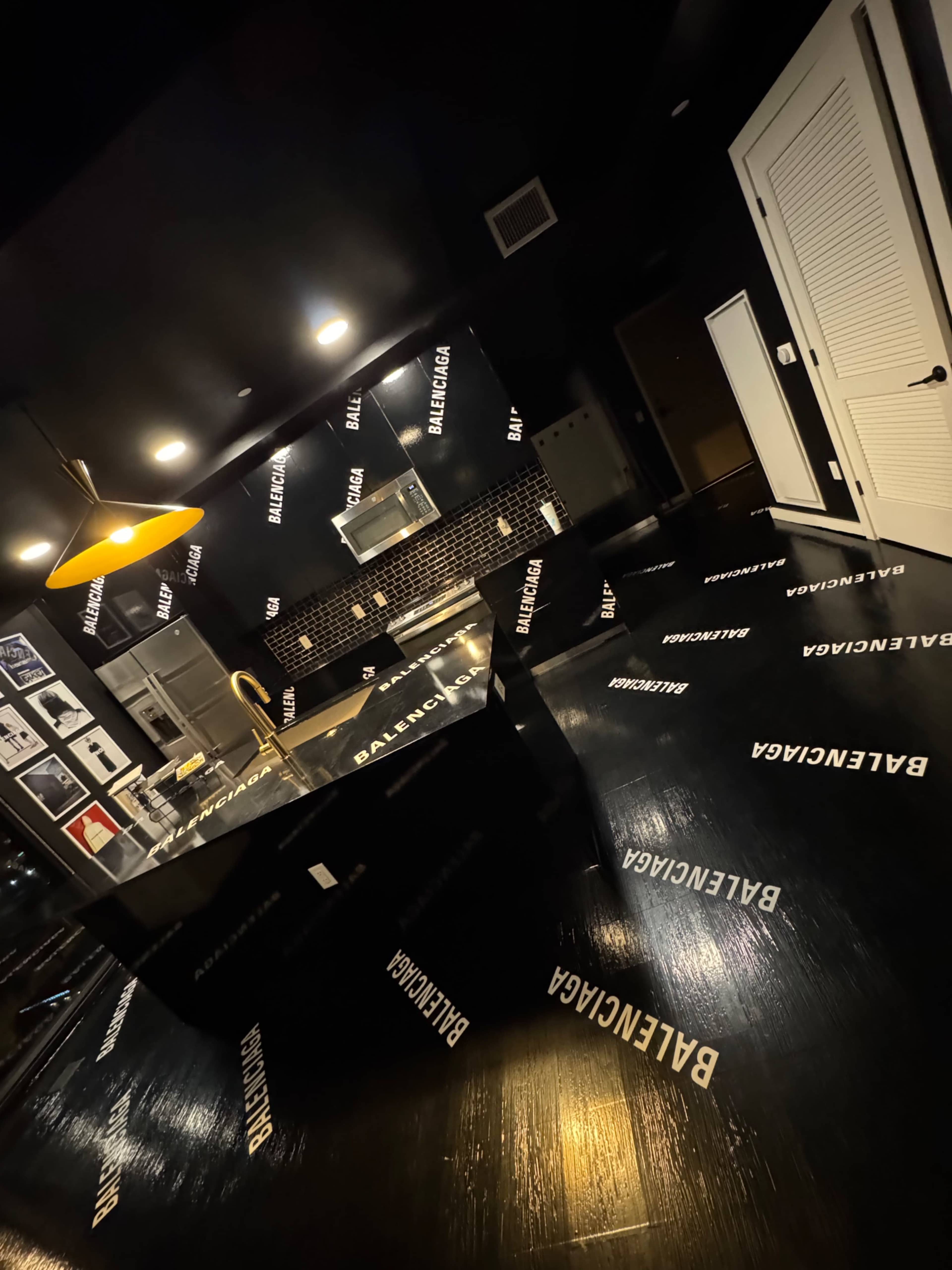 The kitchen features black surfaces adorned with the repeated logo of Balenciaga, illuminated by a hanging light.