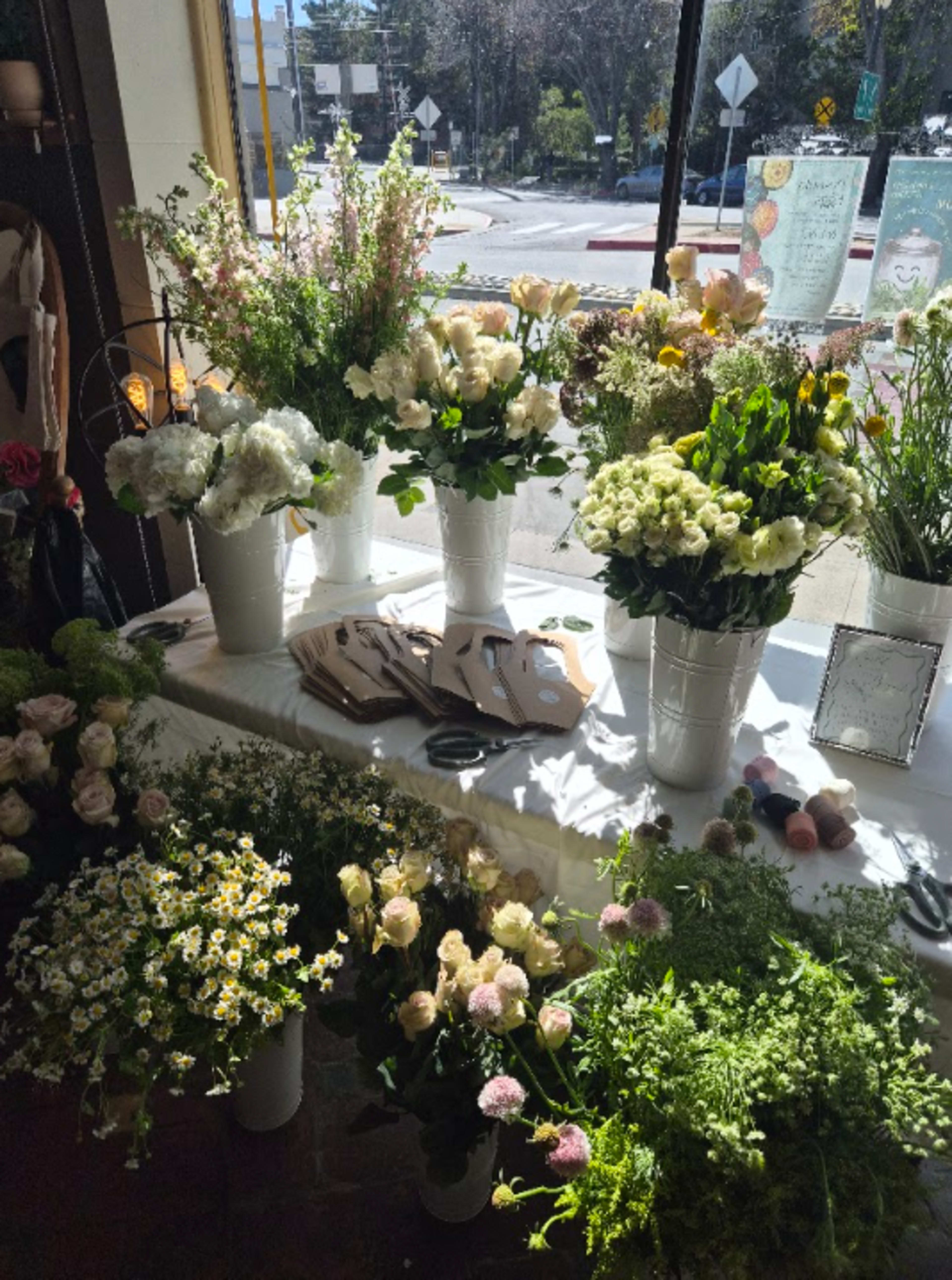 A table filled with various flower arrangements in white pots, surrounded by scattered greenery and tools for floral arrangement.